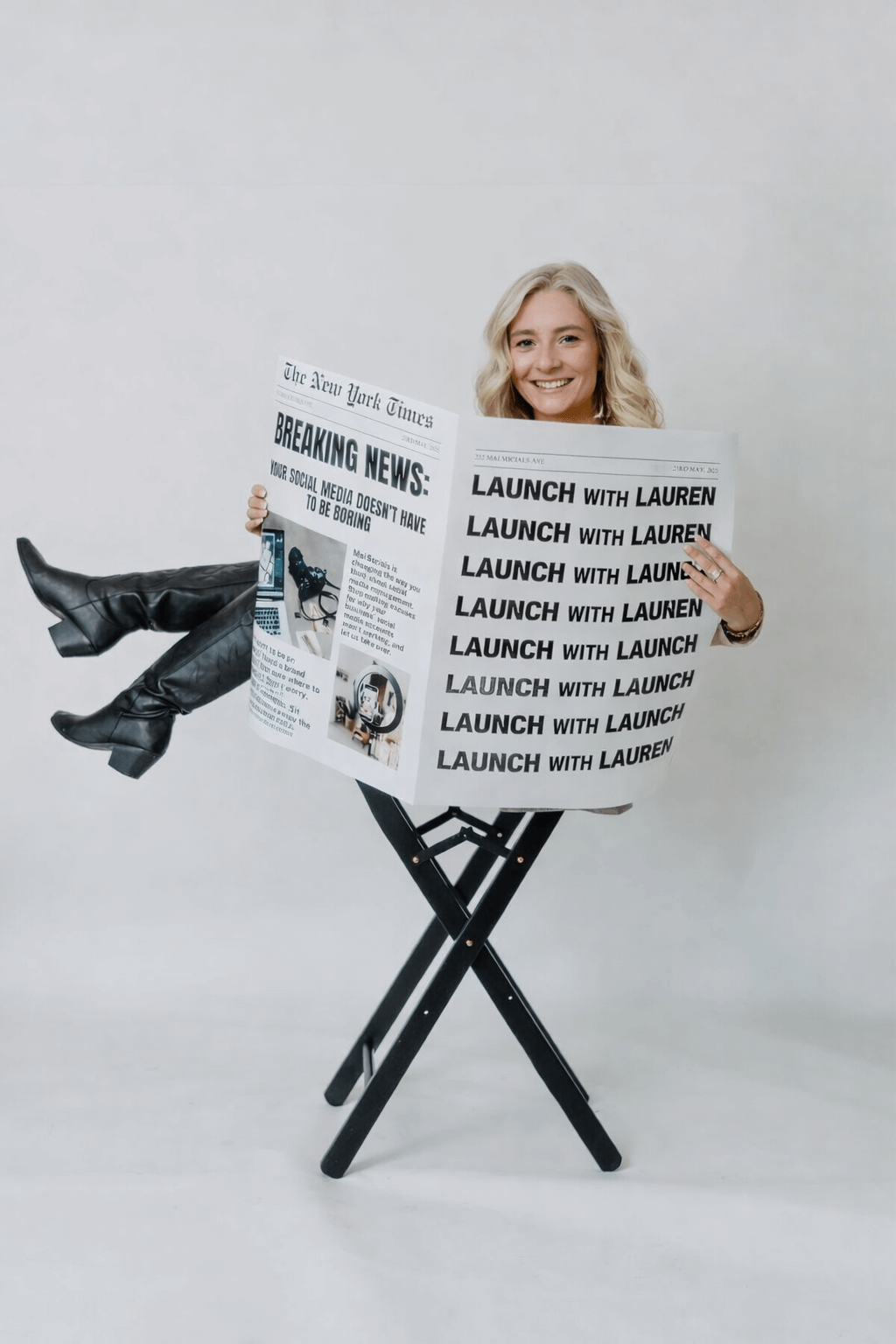 Woman with blonde hair sitting on a director's chair, holding a newspaper with headlines about a launch event, smiling.