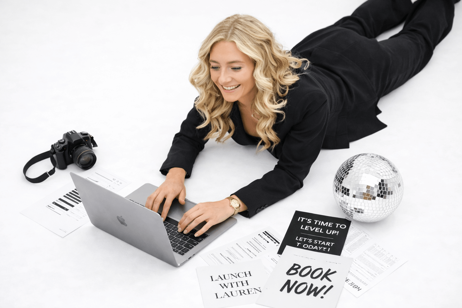 A woman with blonde curly hair laying on the floor using a laptop, surrounded by papers, a camera, a disco ball, and signs that say 'IT'S TIME TO LEVEL UP!', 'LET'S START TODAY!', 'LAUNCH WITH LAUREN', and 'BOOK NOW!'