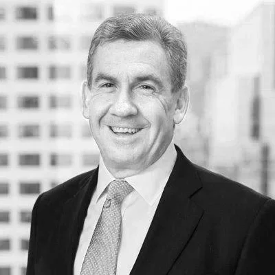 Black and white photo of a smiling man in a suit and tie standing outdoors with a cityscape background.