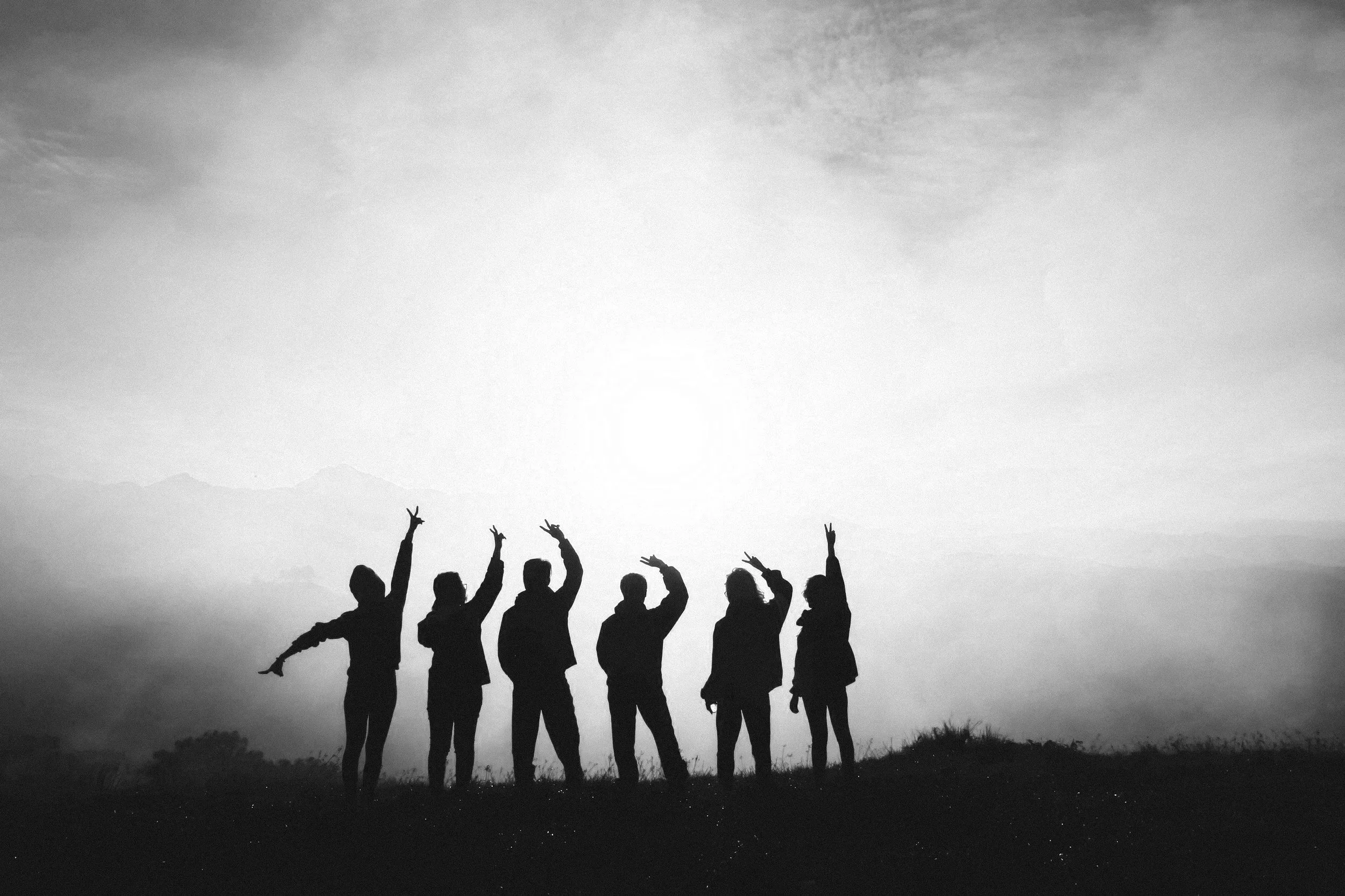 Silhouette of six people standing on a hilltop with a sunset or sunrise in the background, sky with clouds and mountains in the distance.