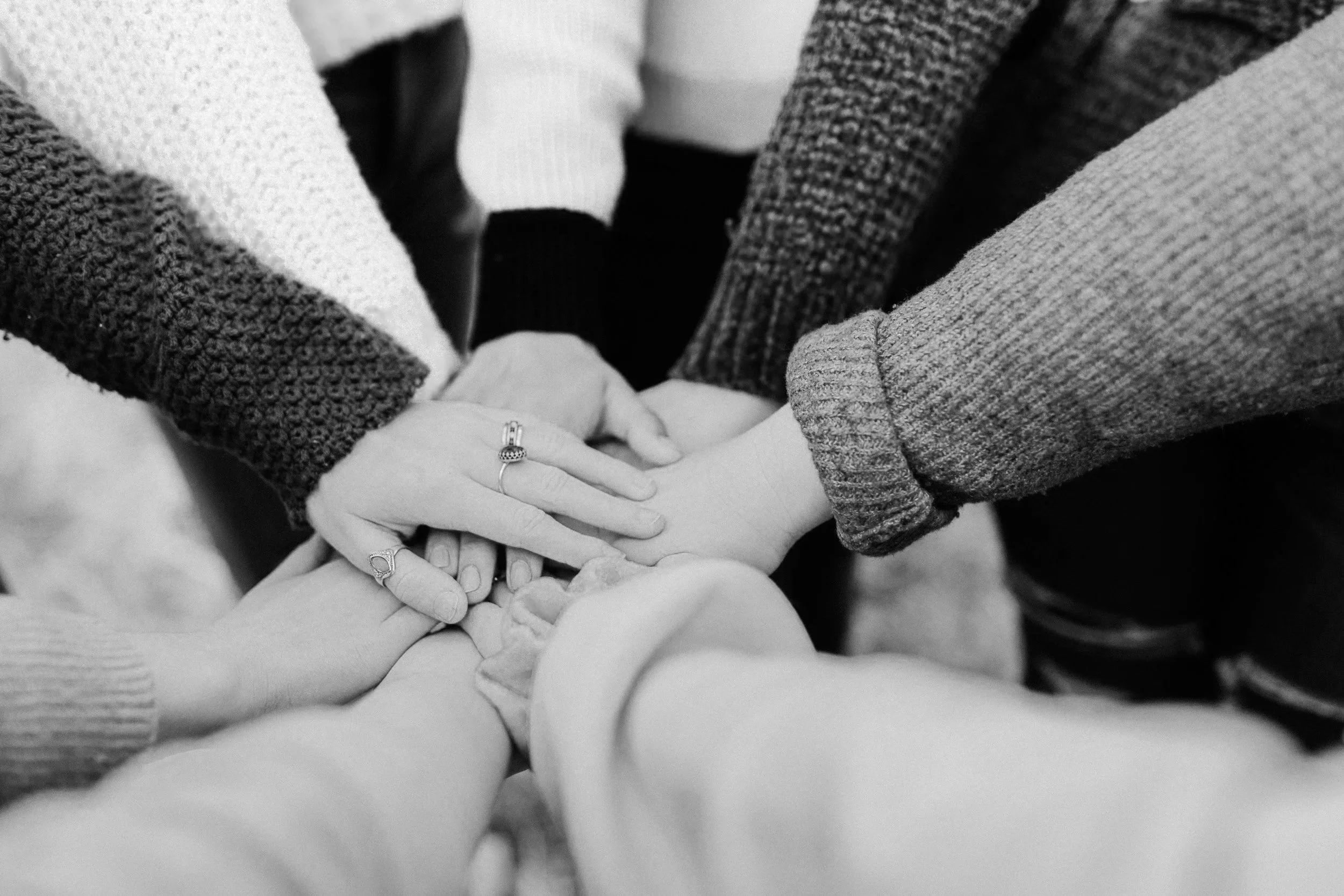 Multiple hands stacked together, some with rings, wearing sweaters and casual clothing, symbolizing unity or teamwork.