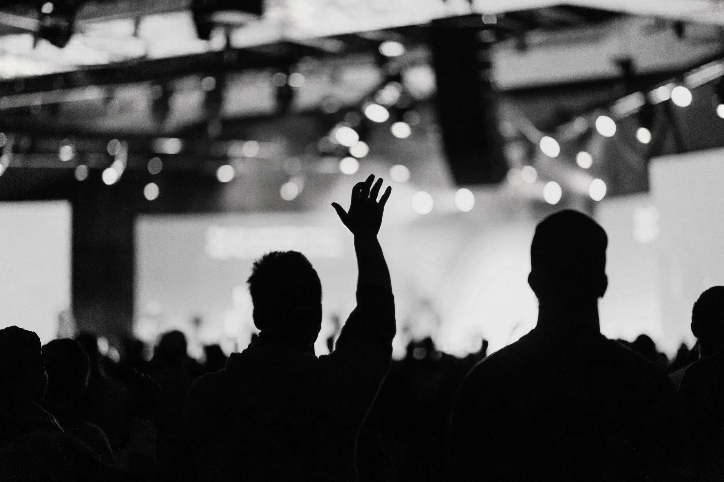 Silhouette of a person raising their hand in a crowd at a concert or event with stage lighting in the background.