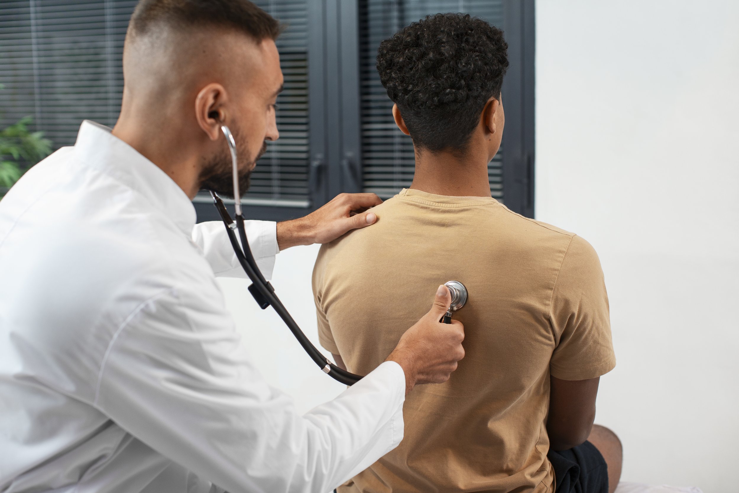 A doctor using a stethoscope to examine a young man's back in a clinic setting.