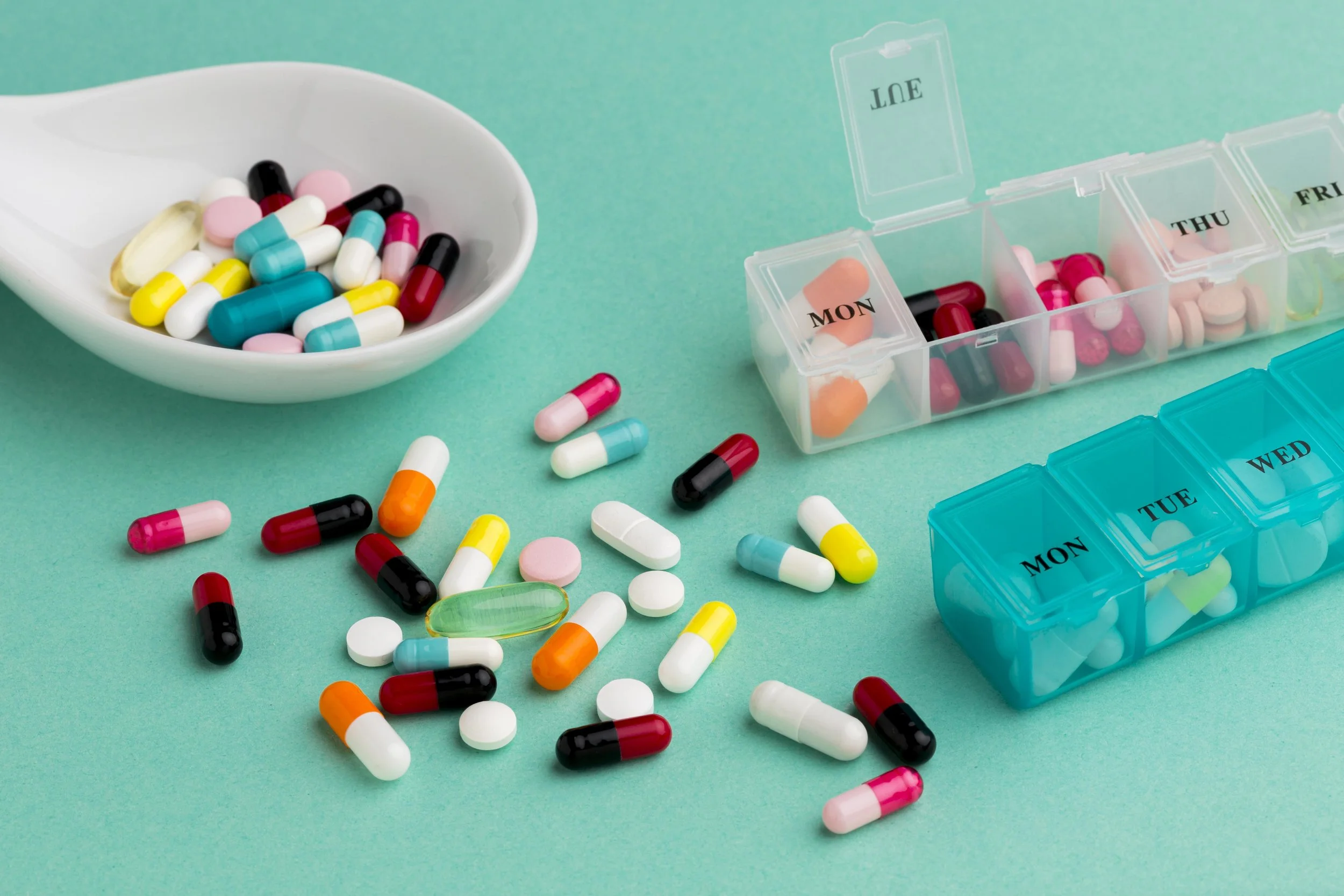 Assorted pills and capsules, some in a white bowl and others scattered on a teal surface, with labeled pill organizers for Monday through Friday.