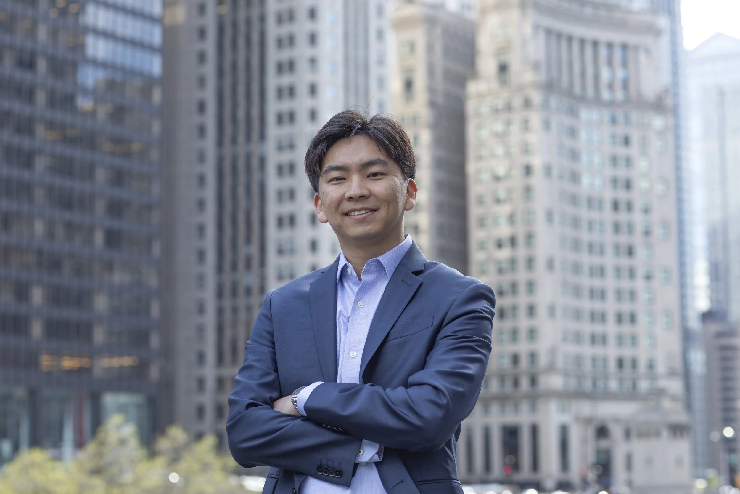 A man in a professional blue suit with crossed arms standing in front of tall city skyscrapers.