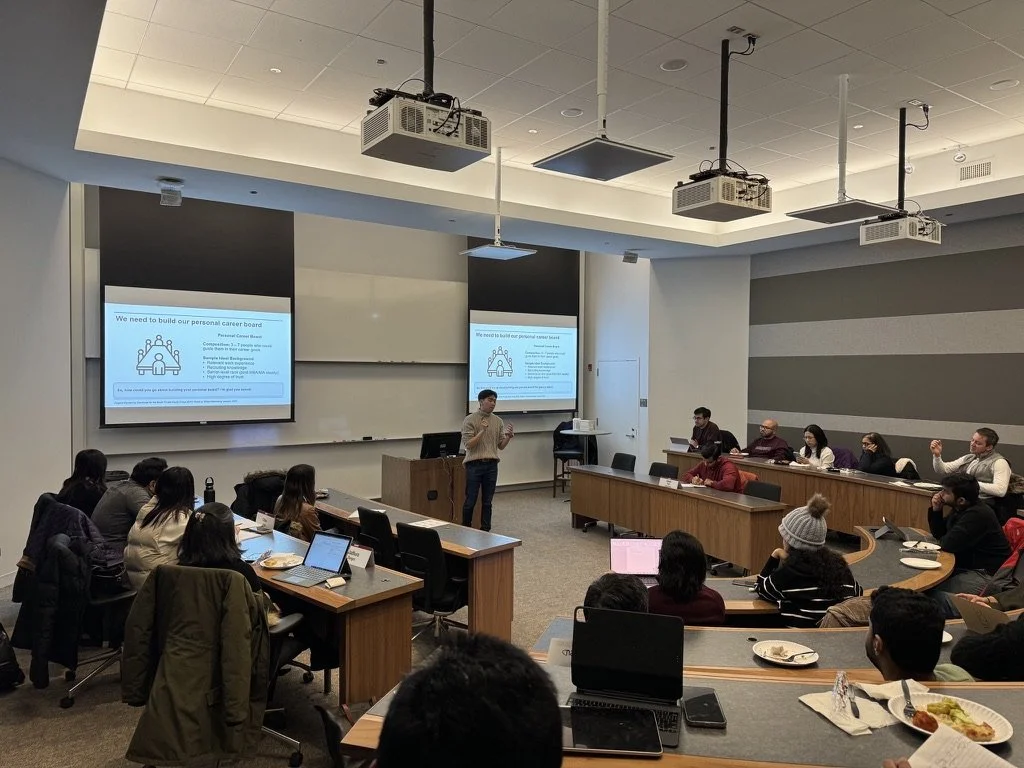 A classroom or conference room with a presenter standing in front of a large screen displaying a slide titled 'We need to build our personal career board.' Students or attendees are seated around tables, some with laptops, taking notes or listening.