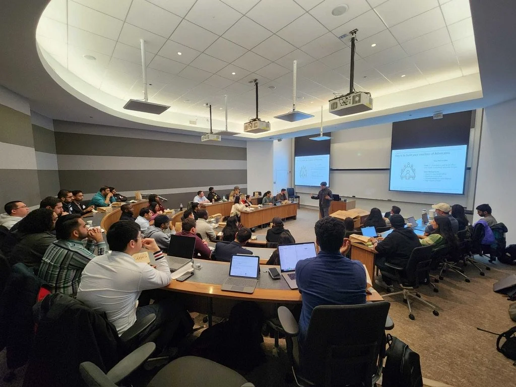 A classroom lecture hall filled with students attention focused on a speaker at the front, using laptops and taking notes.