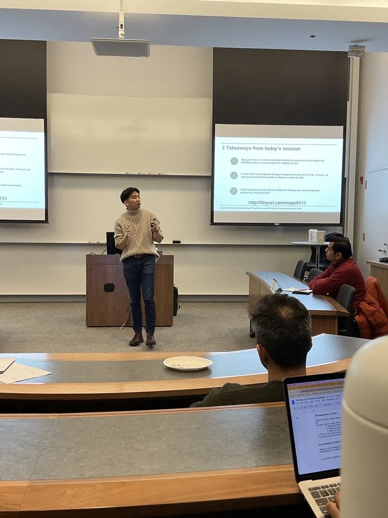 A classroom or lecture hall with a speaker standing at the front, presenting to a group of students. The presentation slide on the screen reads '3 Takeaways from today's session' with bullet points. Several students are seated, some taking notes or working on laptops.