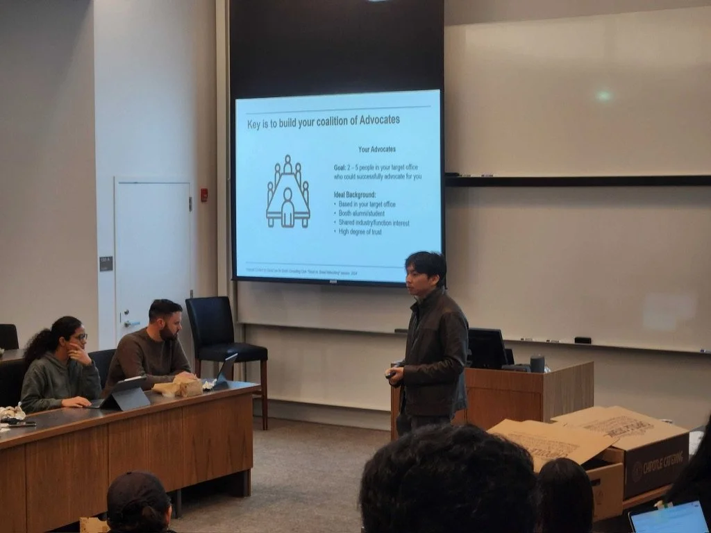 A classroom or conference room with a person giving a presentation. The presentation slide displayed on the large screen reads 'Key is to build your coalition of Advocates' and includes bullet points about advocacy strategies. There are three people seated at a table with laptops and notebooks, and a few audience members in the foreground.