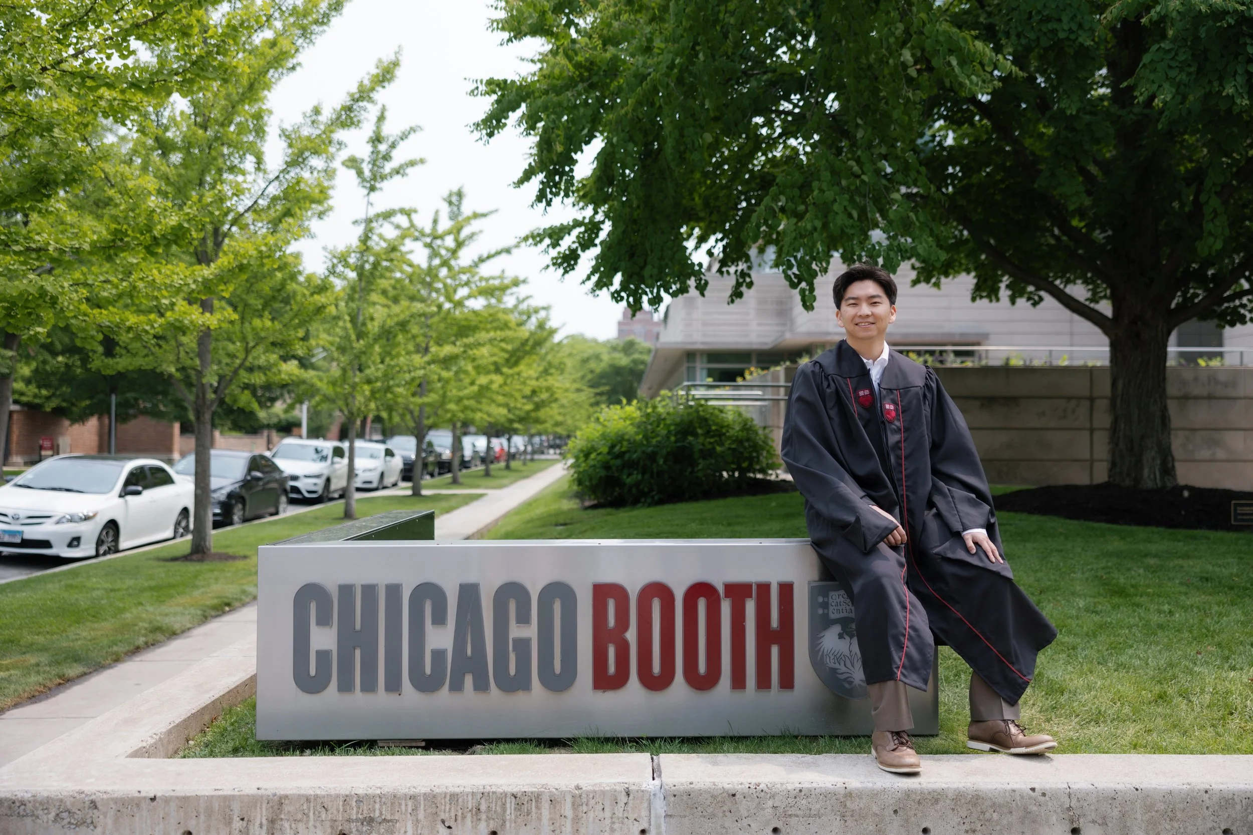 A young man in graduation cap and gown leaning on a sign that reads "Chicago Booth" outside on a college campus with green trees and parked cars in the background.
