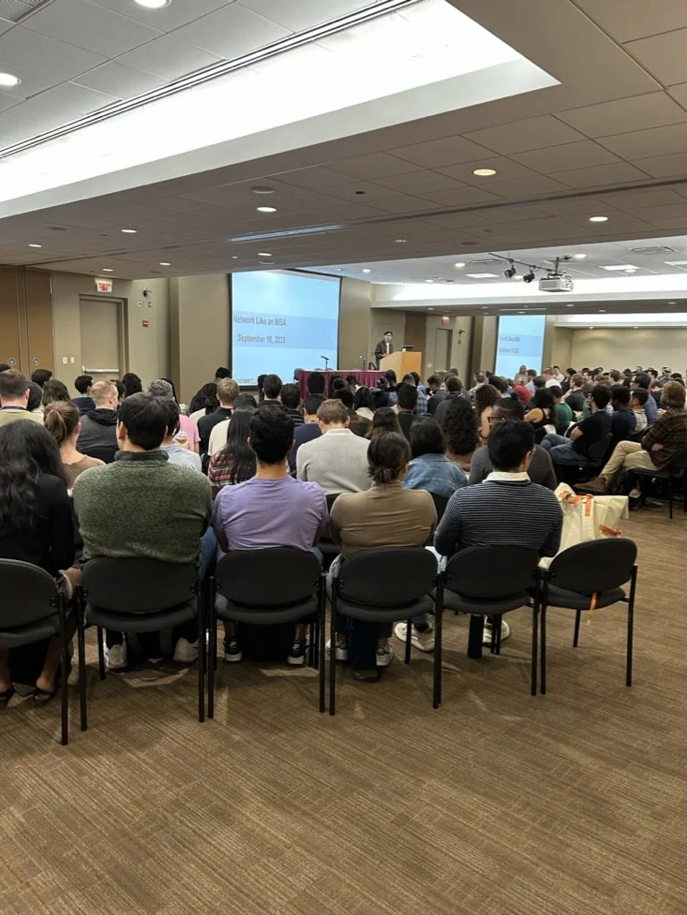 Audience attending a conference or seminar in a large room, with a person speaking at a podium in front of a projected screen that reads 'Network Like an MSBA, September 11, 2023.'
