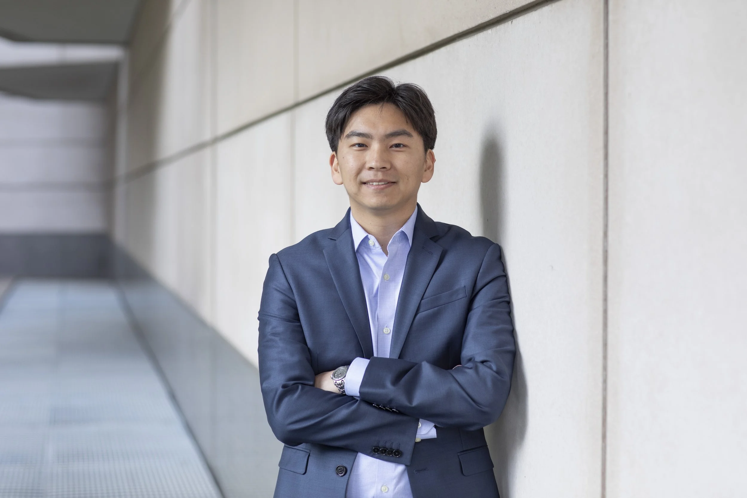 Young man in a blue suit smiling with arms crossed, standing against a concrete wall outdoors.
