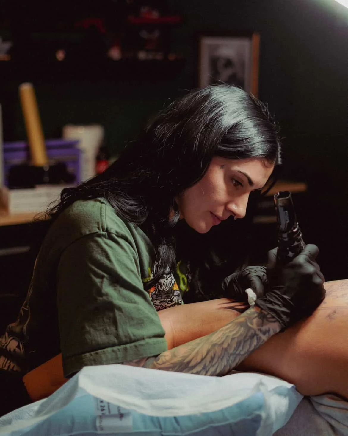A woman with long black hair, wearing black gloves and a green shirt, is tattooing the leg of a person lying on a table in a tattoo studio.