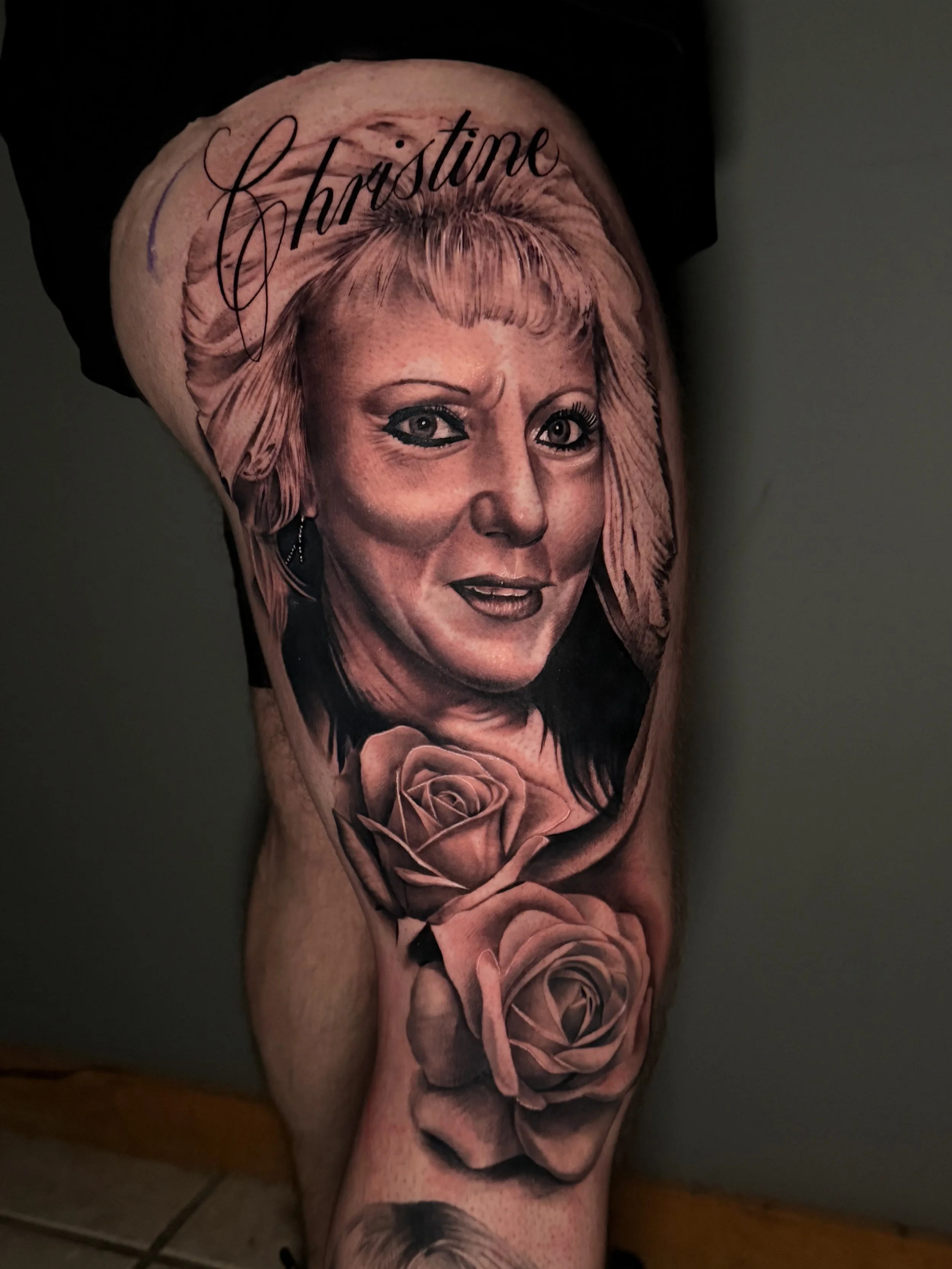 Realistic black and gray tattoo of a woman's face with the name 'Christine' written above, surrounded by two detailed roses on the lower part of the leg.