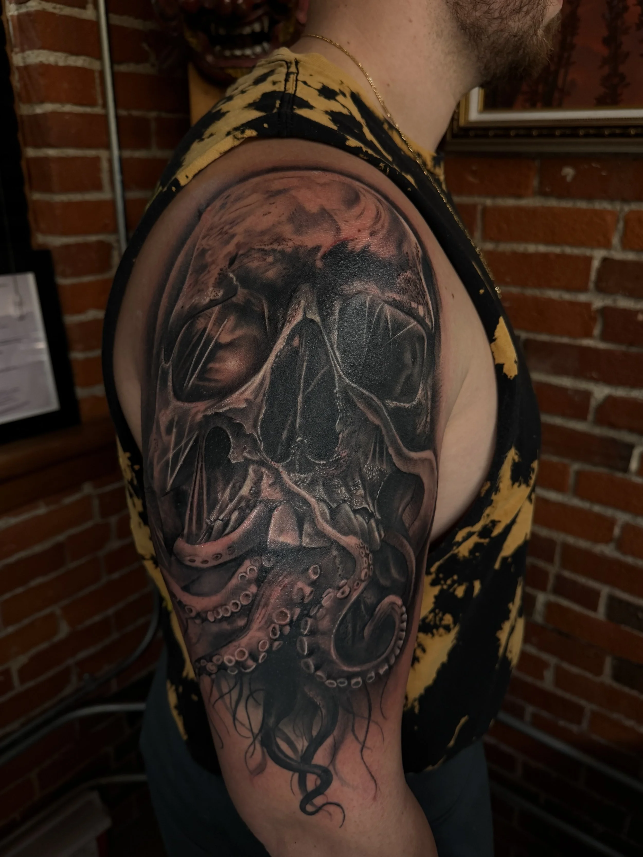 Tattoo of a skull with an octopus tentacle extending from the jaw on a man's upper arm and shoulder.