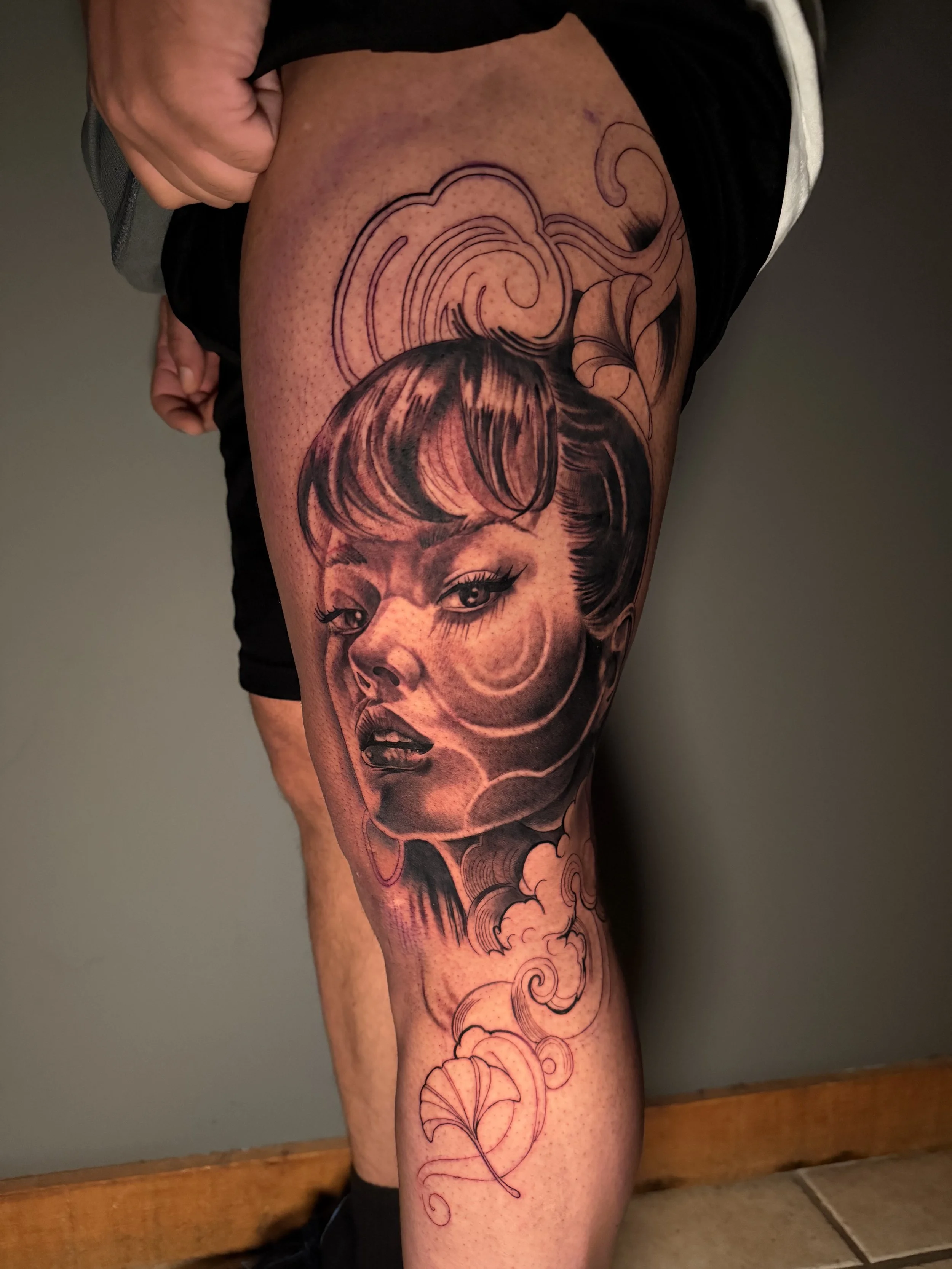 A detailed black and gray portrait tattoo of a woman with bangs and hoop earrings on a person's thigh, surrounded by swirling clouds and floral designs.