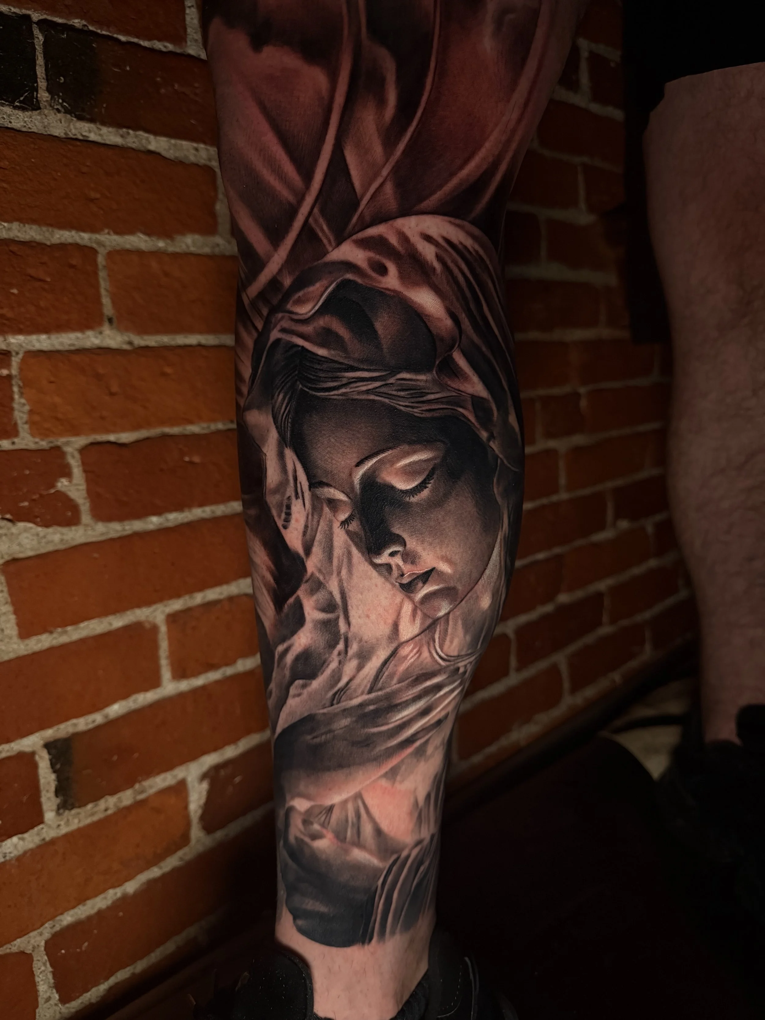 Black and gray tattoo of a serene woman with a hooded cloak, closed eyes, and delicate features on a person's leg, set against a brick wall background.