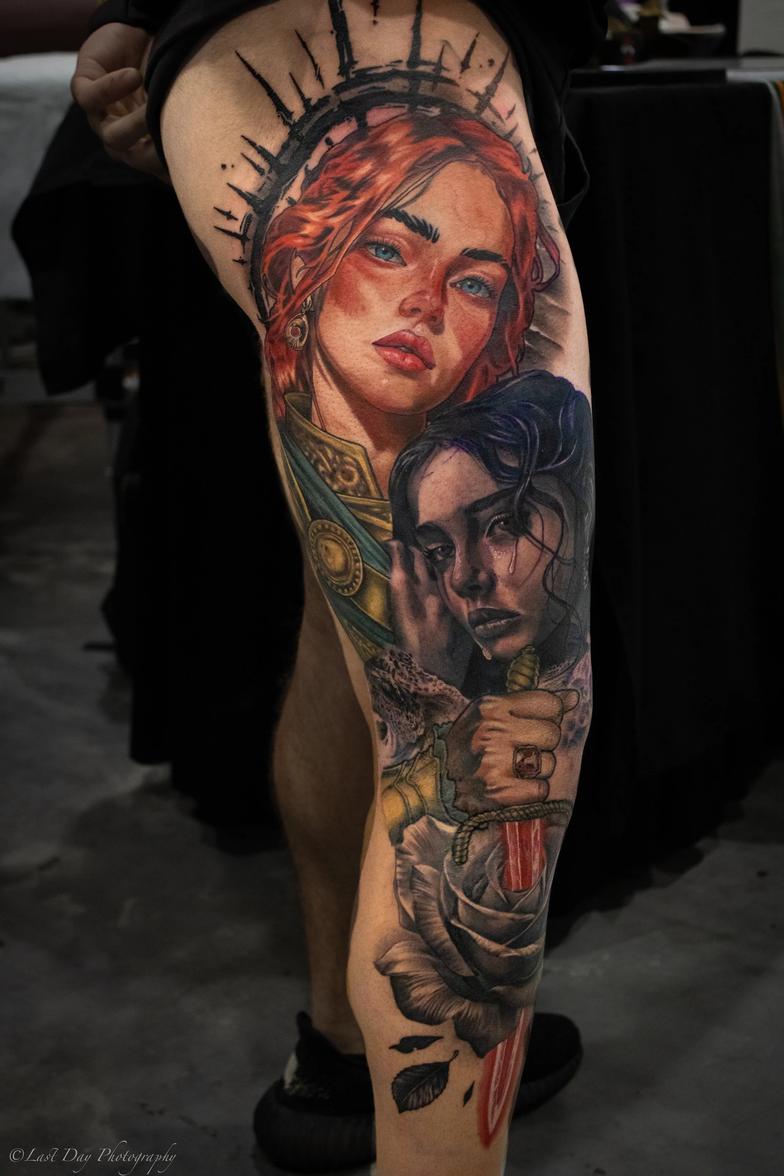 A detailed colorful tattoo sleeve featuring two women. The top woman has red hair, blue eyes, and a halo-like black symbol above her head. The bottom woman has dark wavy hair and is holding a dagger wrapped in cloth, with a snake and leaves near her 
