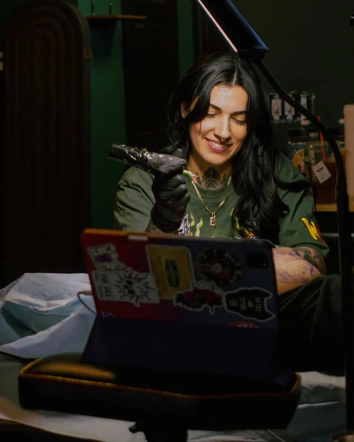 A woman with black hair, tattoos, and a septum piercing is smiling while sitting at a table. She is holding a tattoo machine in her right hand, wears black gloves, a green shirt with patches, and a necklace. There is a tablet with stickers on the table, along with a white paper towel, and a bag in the background.