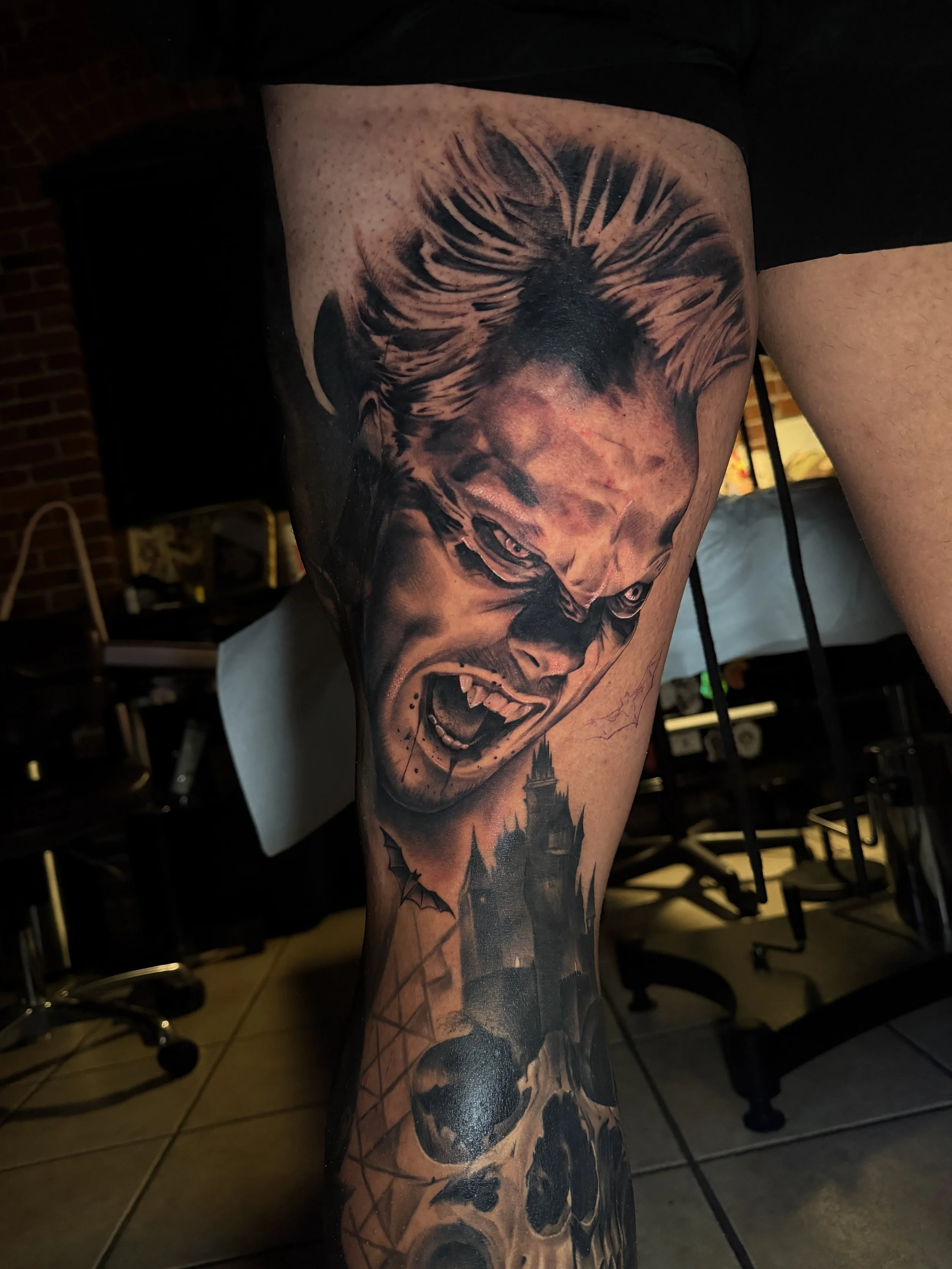 A black and gray tattoo on a person's thigh depicting a screaming face with wild hair and fanged teeth, with a haunted house and skulls below.