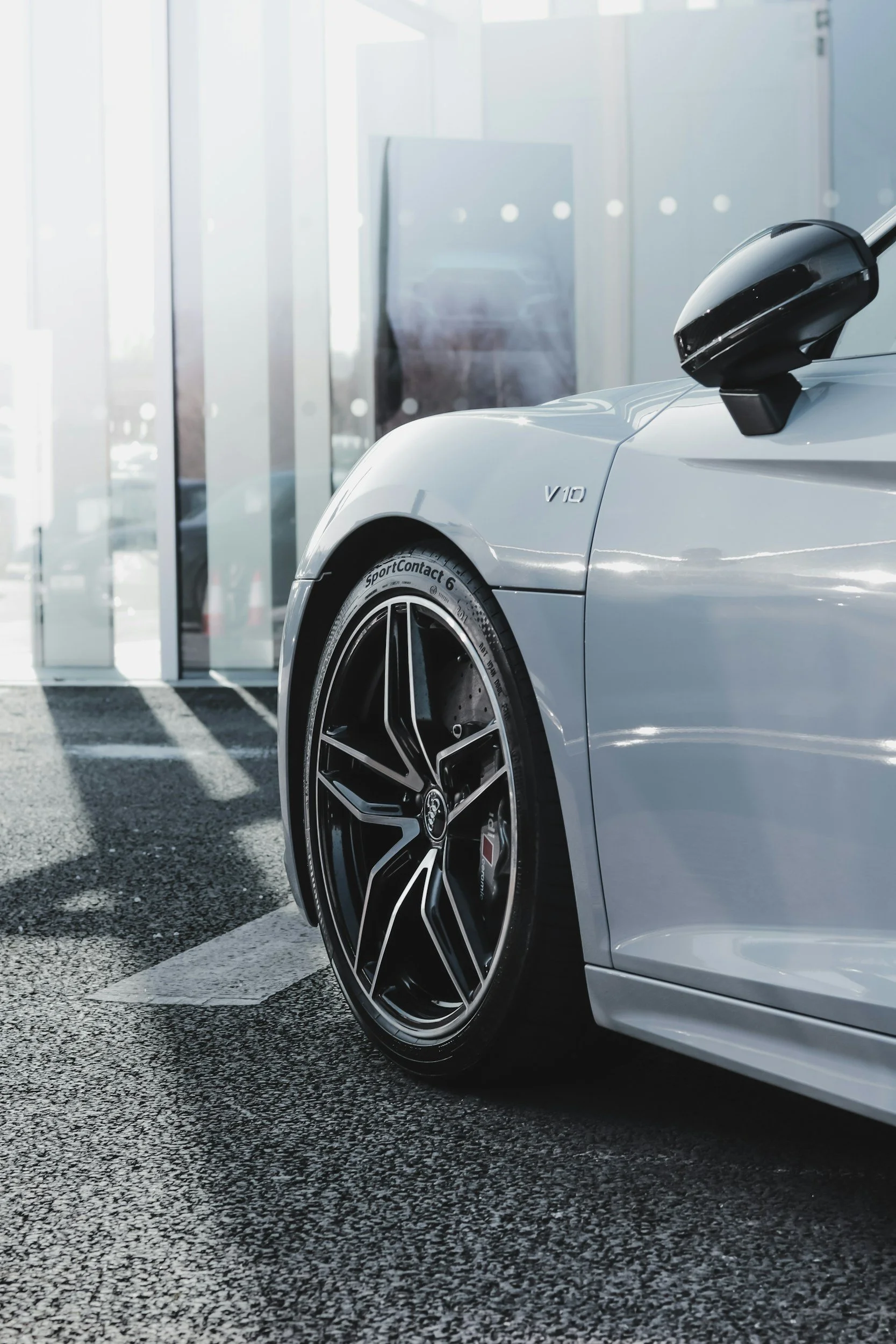 Close-up of a silver luxury sports car showing front left tire and part of the door, with sunlight casting shadows on the pavement outside a modern building.