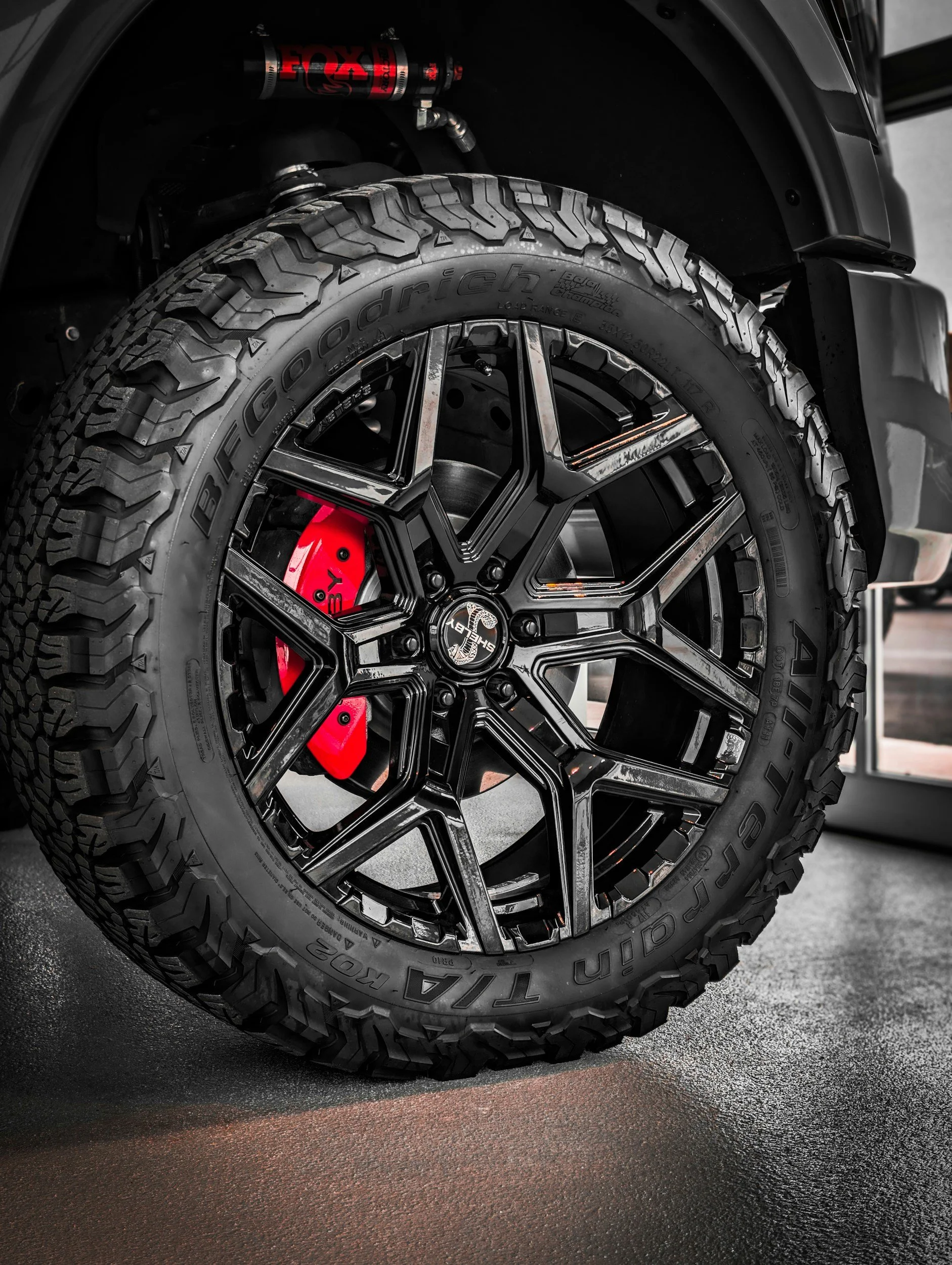 Close-up of a vehicle's tire with a large off-road tread pattern, mounted on a black alloy wheel with multiple spokes. The brake caliper behind the wheel is painted red.