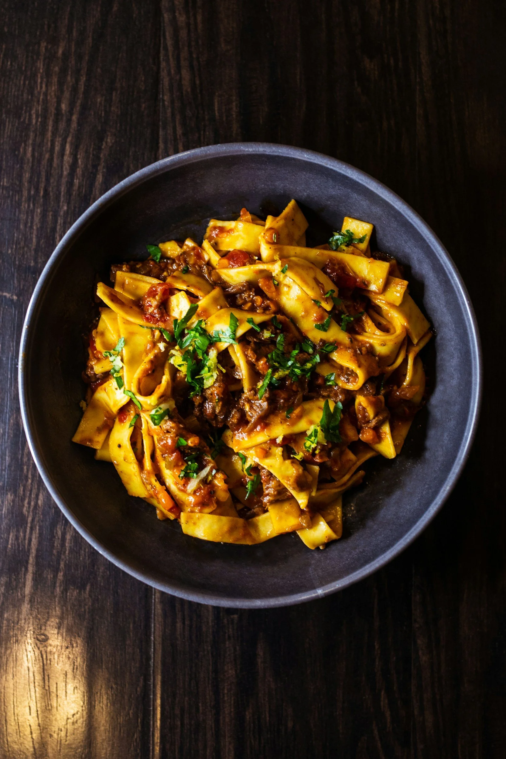 A bowl of pasta topped with sauce, herbs, and vegetables on a dark wooden surface.