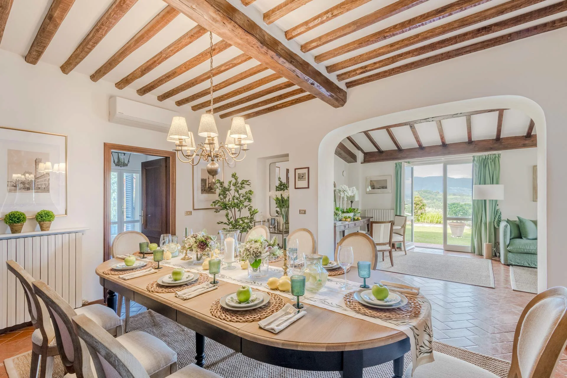 Dining room with a wooden table set for a meal, surrounded by beige chairs, with a chandelier overhead. The room has white walls, wooden beams on the ceiling, and large windows showing a scenic outdoor view of green hills.