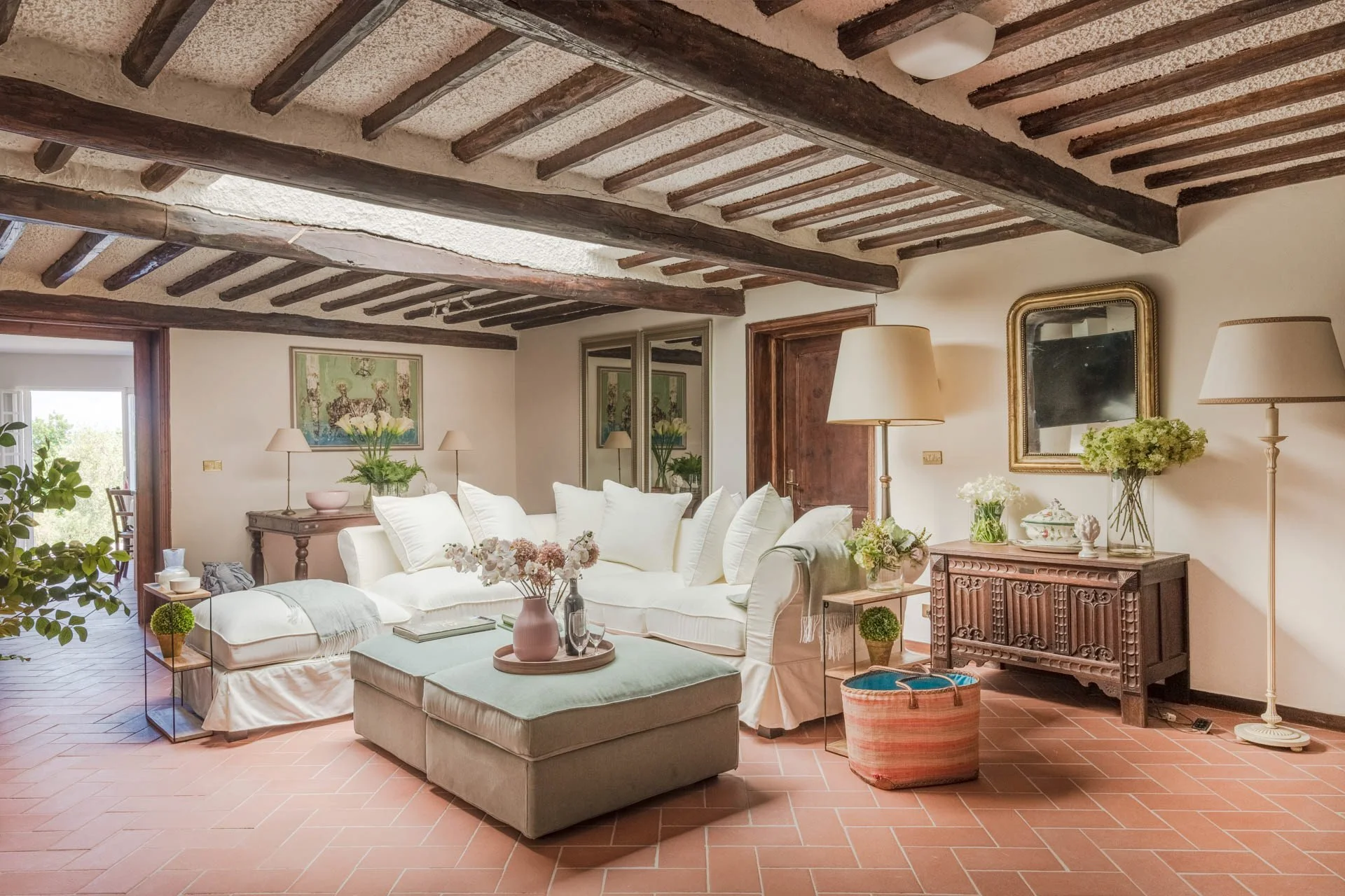 Living room with white sofa, green ottoman, wooden beams on ceiling, floor lamps, potted plants, framed artwork, mirror, and door leading to balcony.