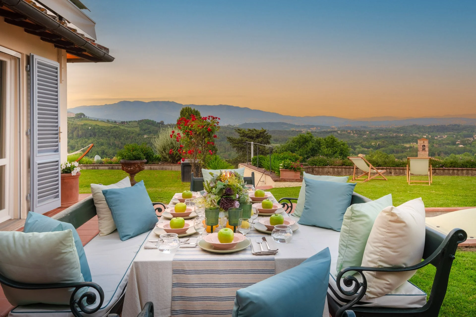 Outdoor dining table set on a patio with plates, glasses, green apples, and a floral centerpiece, overlooking a green lawn and scenic mountains at sunset.