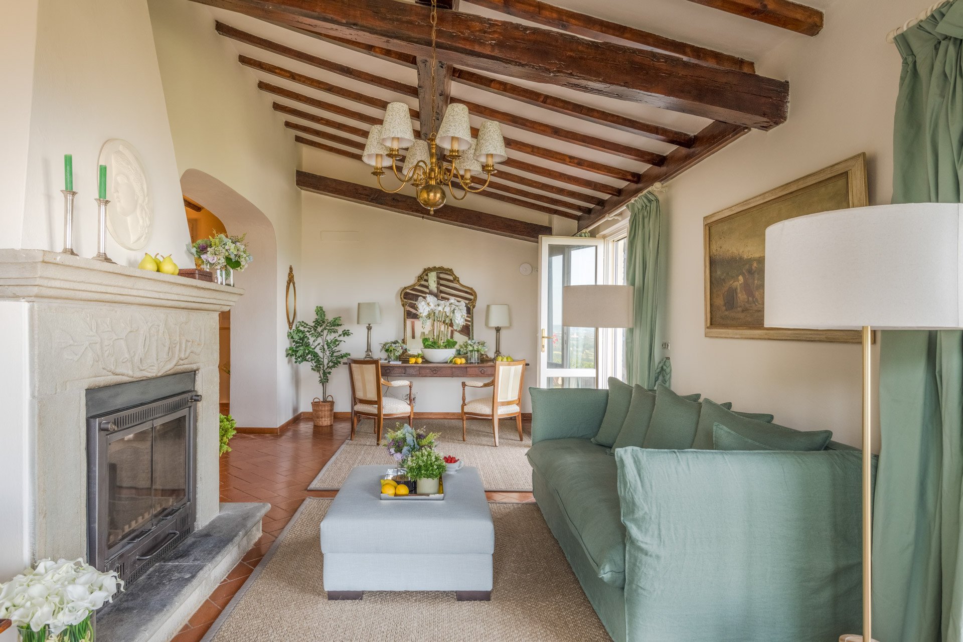 Interior of a living room with a vaulted ceiling featuring exposed wooden beams, a large window with green curtains, a green sofa with matching pillows, a floor lamp, a fireplace with decorative items on mantel, a wooden dining table with chairs, and