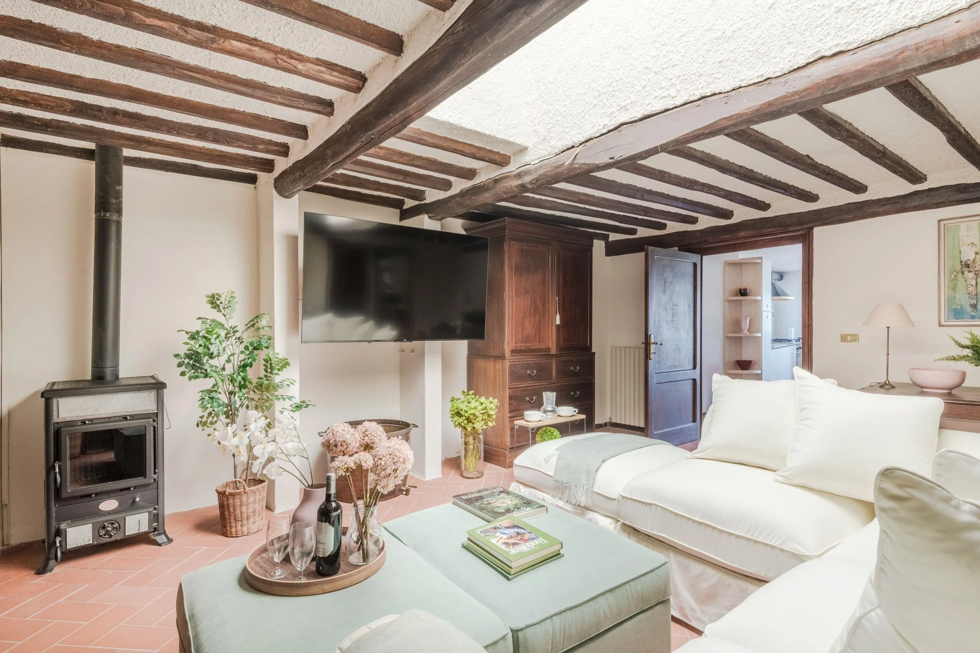 Living room with white sofas, a wooden cabinet, potted plants, a flat-screen TV, and a wood stove. Exposed wooden beams on the ceiling and a door to another room.