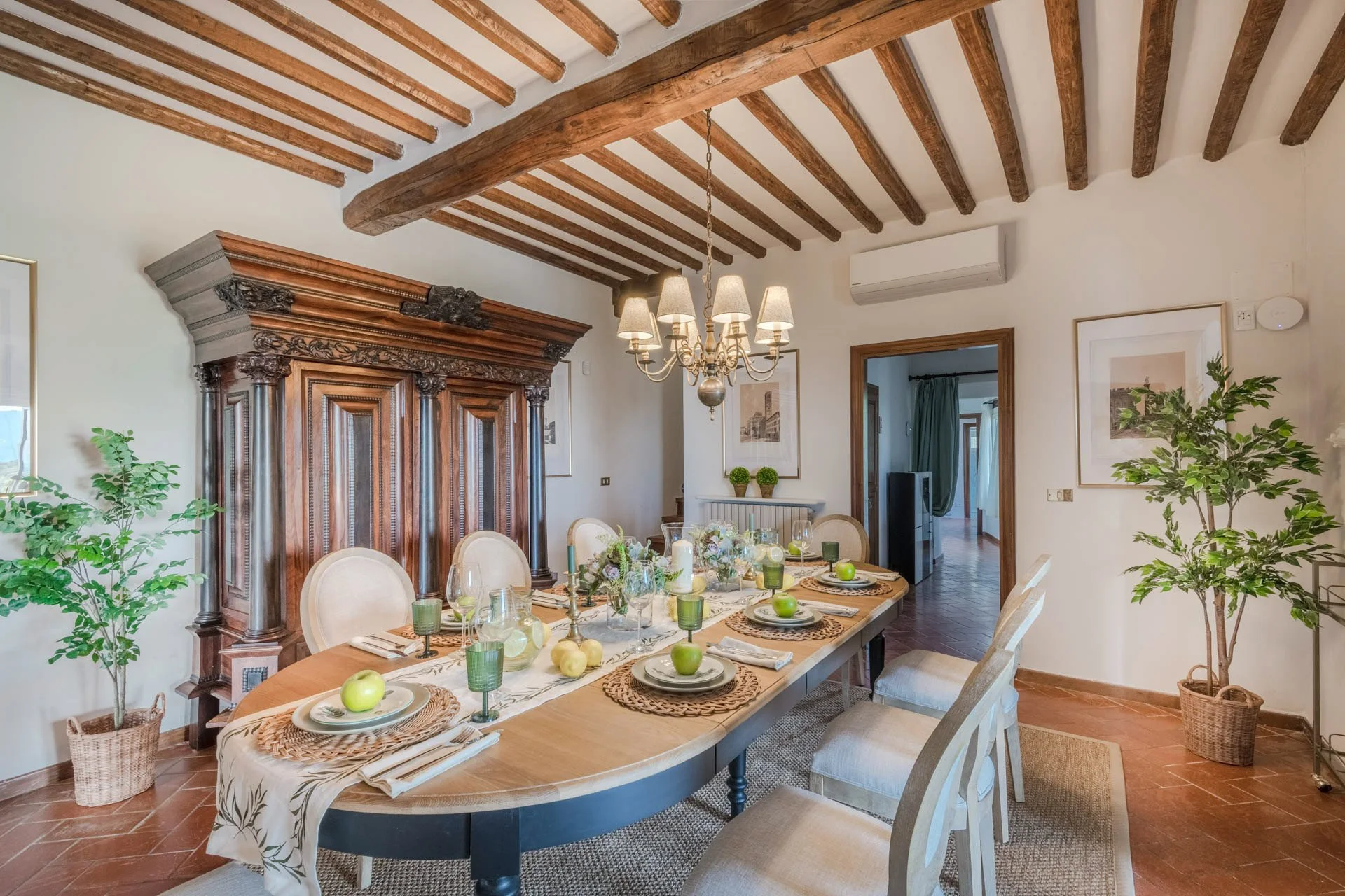 A dining room with a large wooden table set with plates, apples, candles, and glassware, surrounded by beige upholstered chairs. The room features a vintage wooden armoire, wooden ceiling beams, a chandelier, potted plants, framed artwork, and walls 