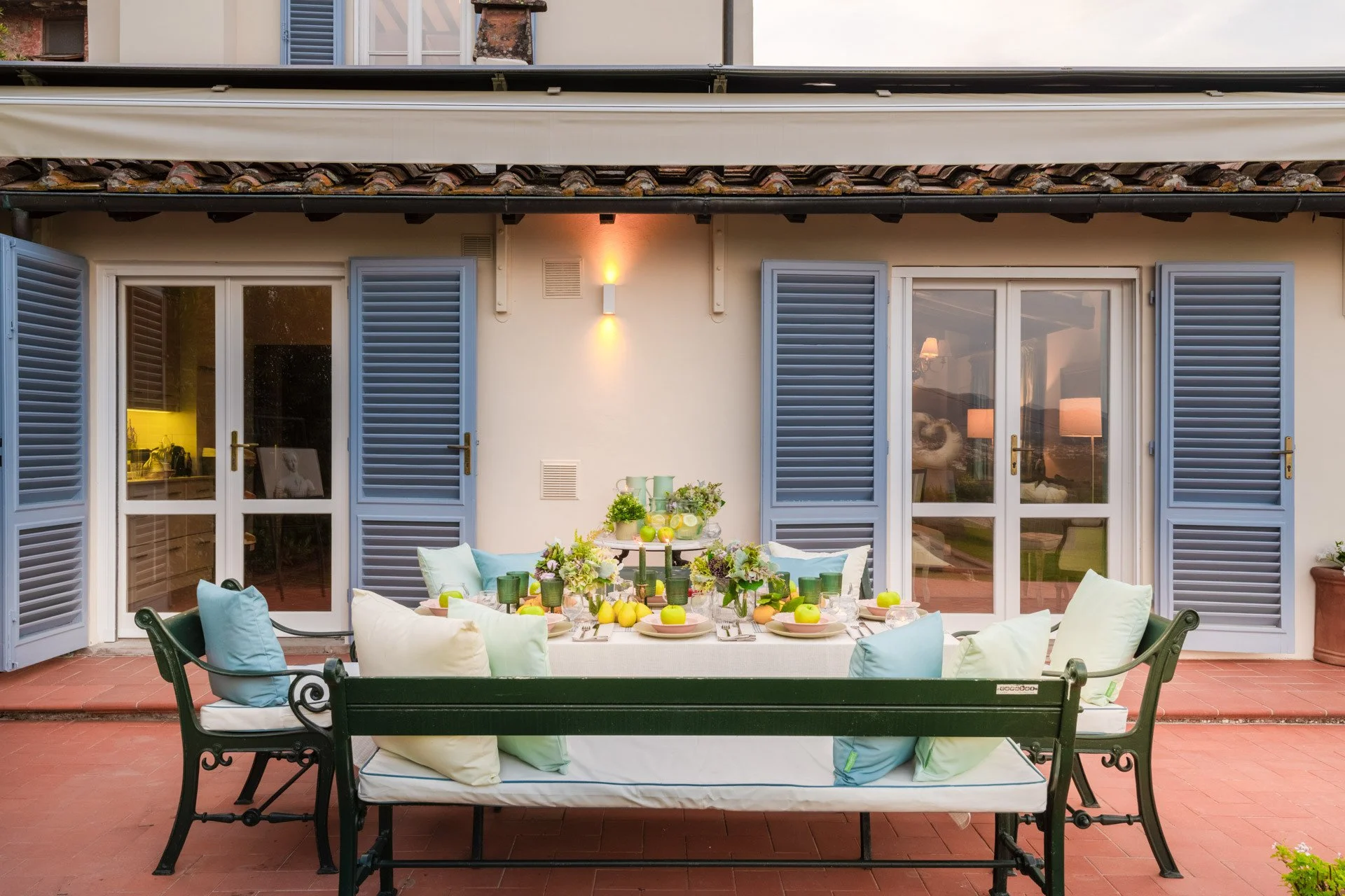 An outdoor dining table set up on a patio with cushions, surrounded by blue shutters and glass doors, decorated with green candles, lemons, and flowers, with warm lighting inside.