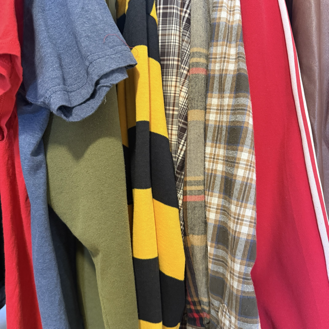 Clothes hanging on a rack, including a gray T-shirt, a blue denim shirt, a green sweater, a black and yellow striped scarf, a plaid shirt, a gray cardigan, and a red piece of clothing.