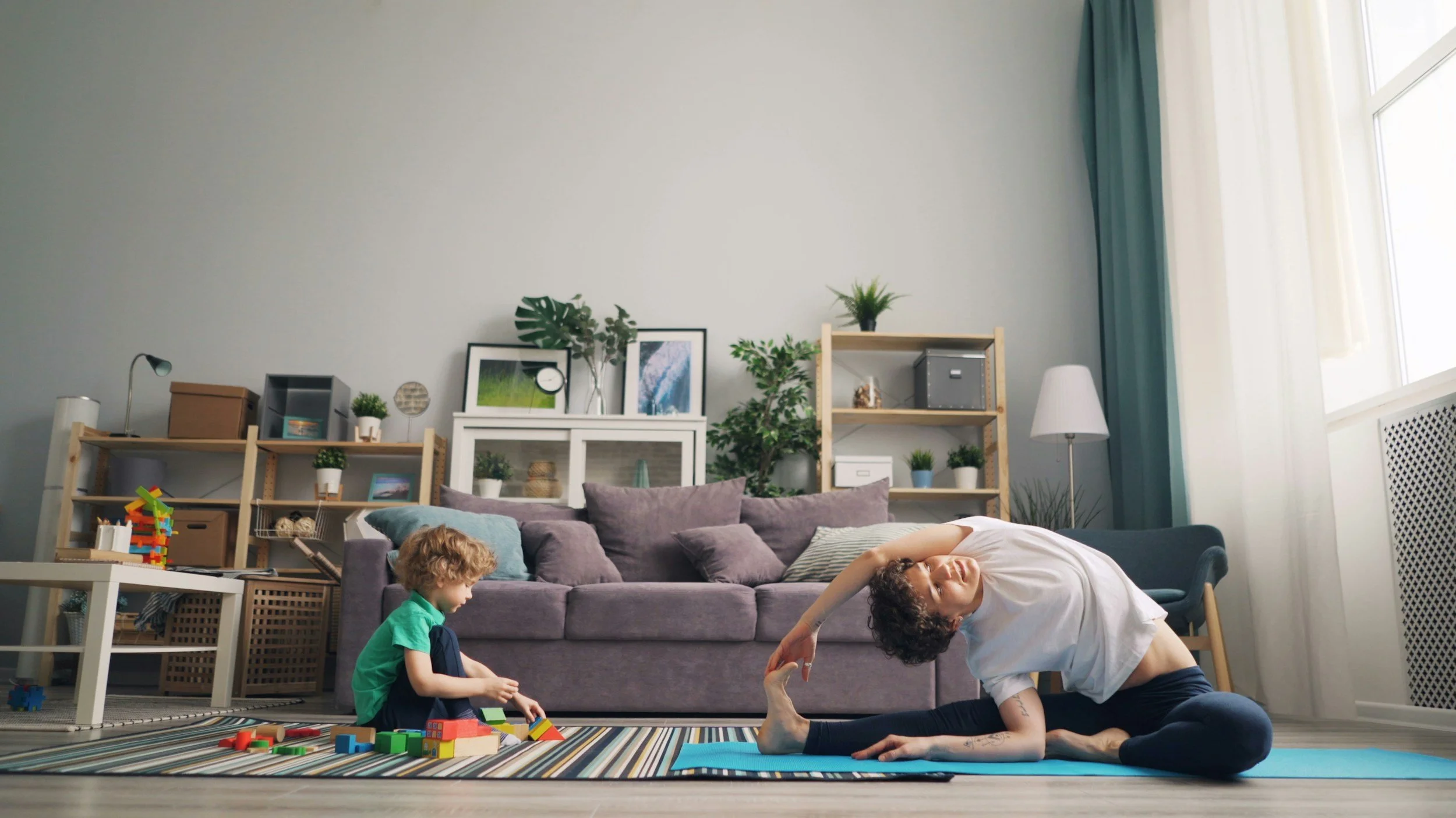 A woman practicing yoga in a living room while a young boy plays with colorful building blocks nearby.