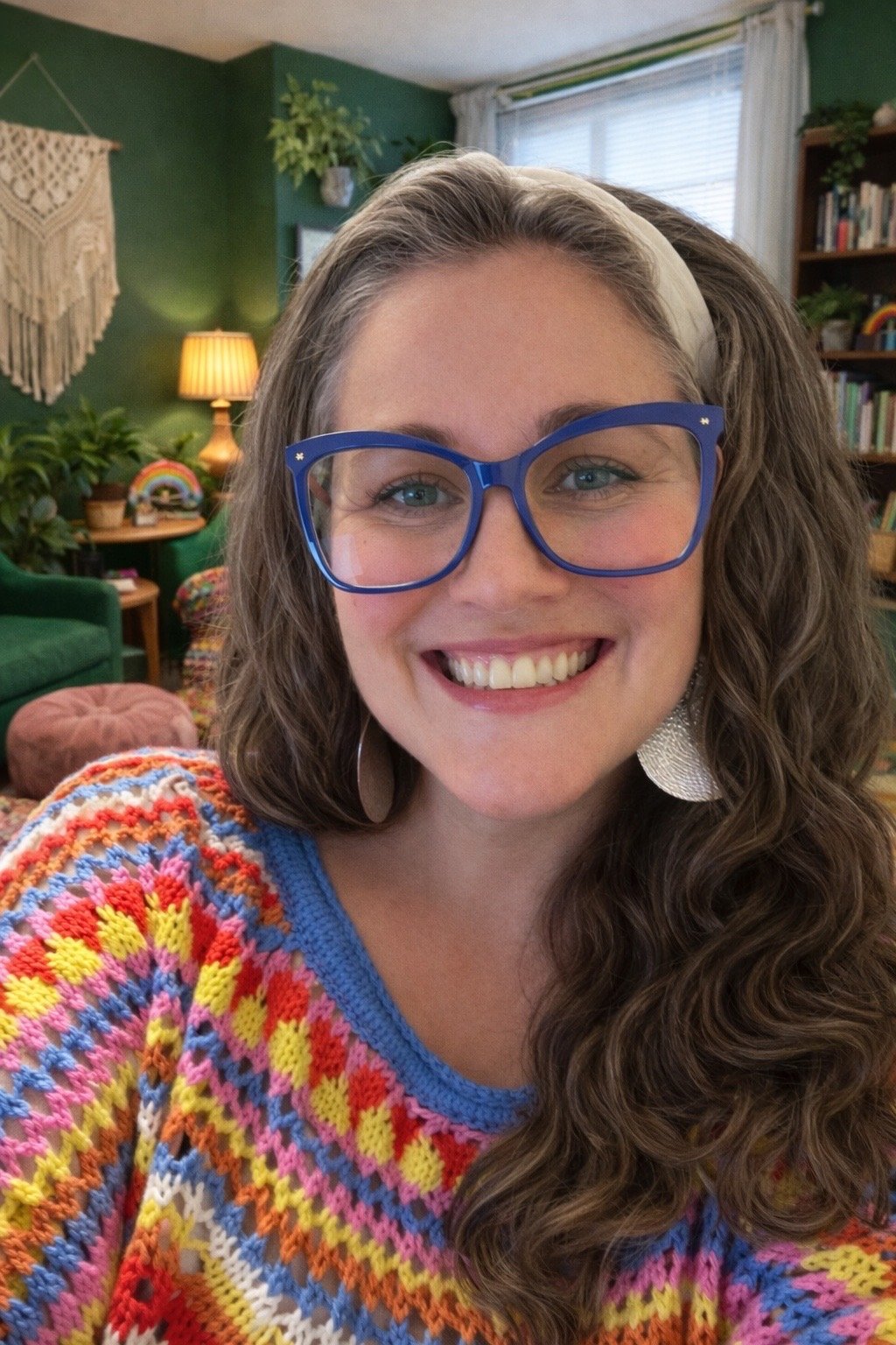 A woman with curly brown hair, wearing blue glasses, smiling broadly, in a cozy living room with green walls, a bookshelf, a lamp, plants, and colorful decorations.