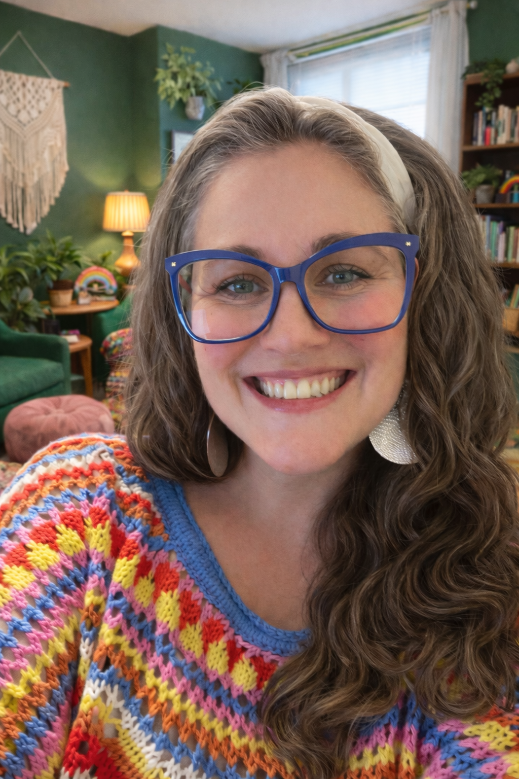 A woman with long curly hair, blue glasses, and a white headband smiling in a cozy living room with green walls, plants, a bookshelf, and colorful decorations.