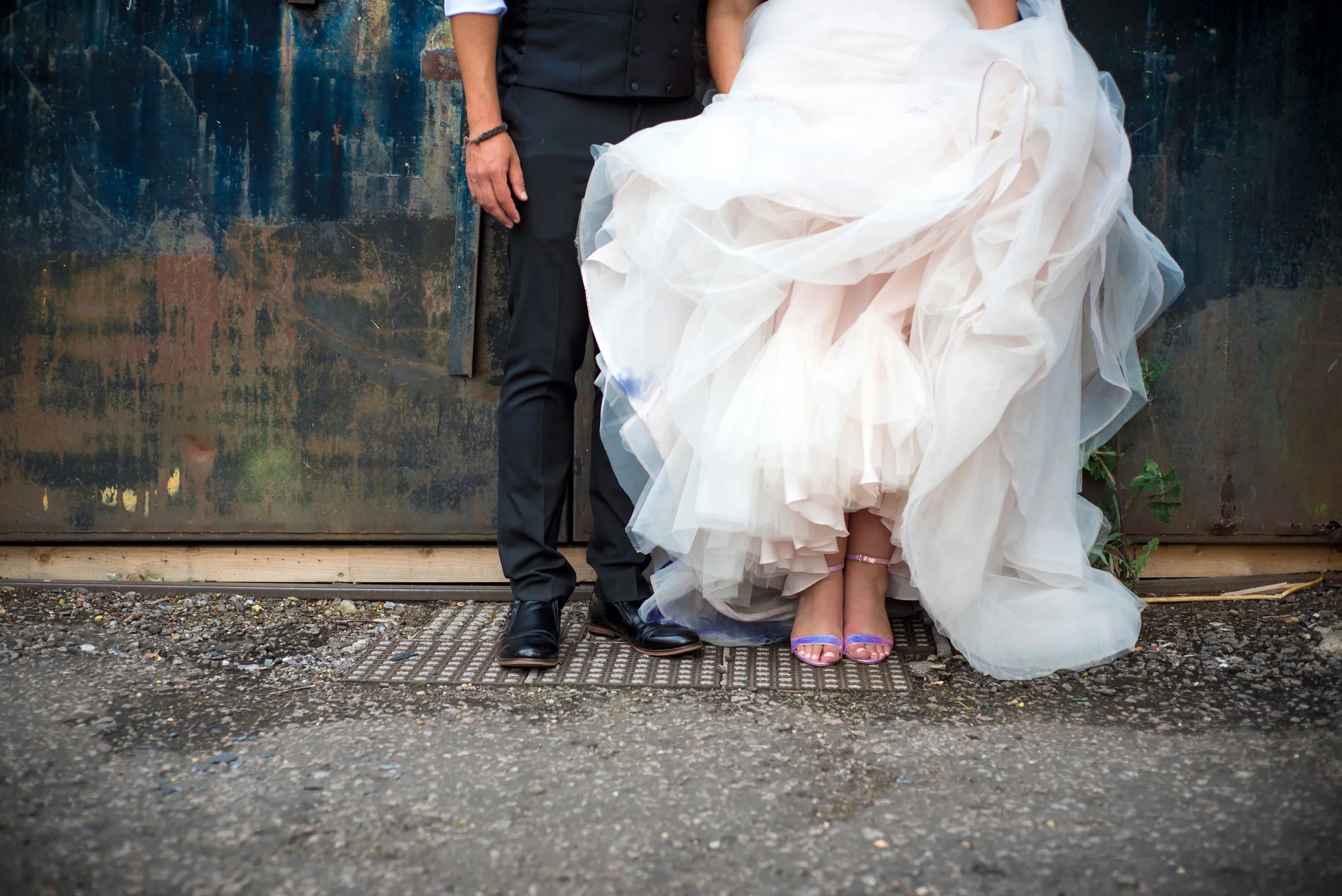 Close-up of a bride in a wedding gown and heels, and a groom in a black suit and shoes, standing side by side outdoors on a gravel surface, with a rusty metal background.