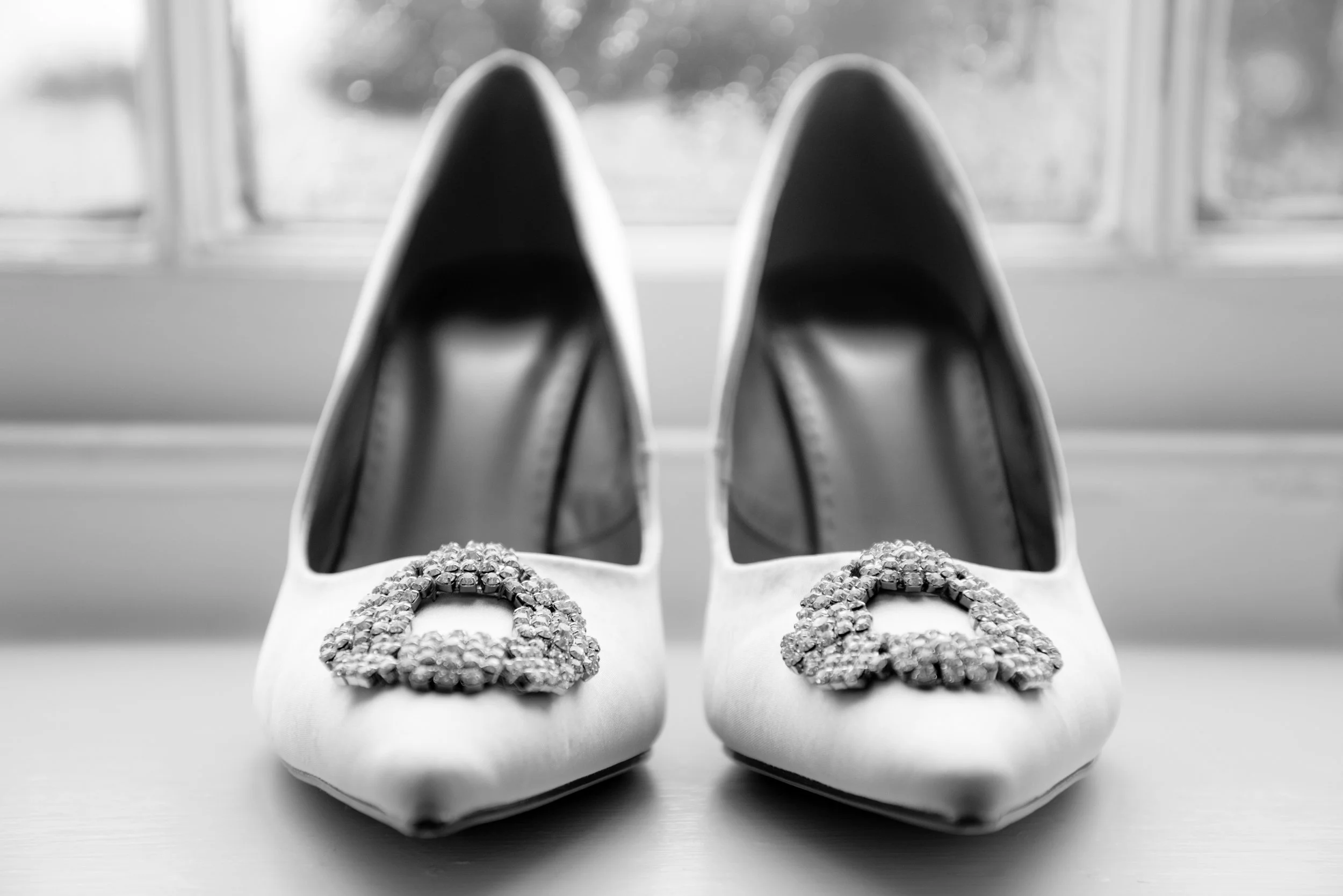 A pair of white high-heeled shoes with rhinestone embellishments on the toes, placed on a surface in front of a window.