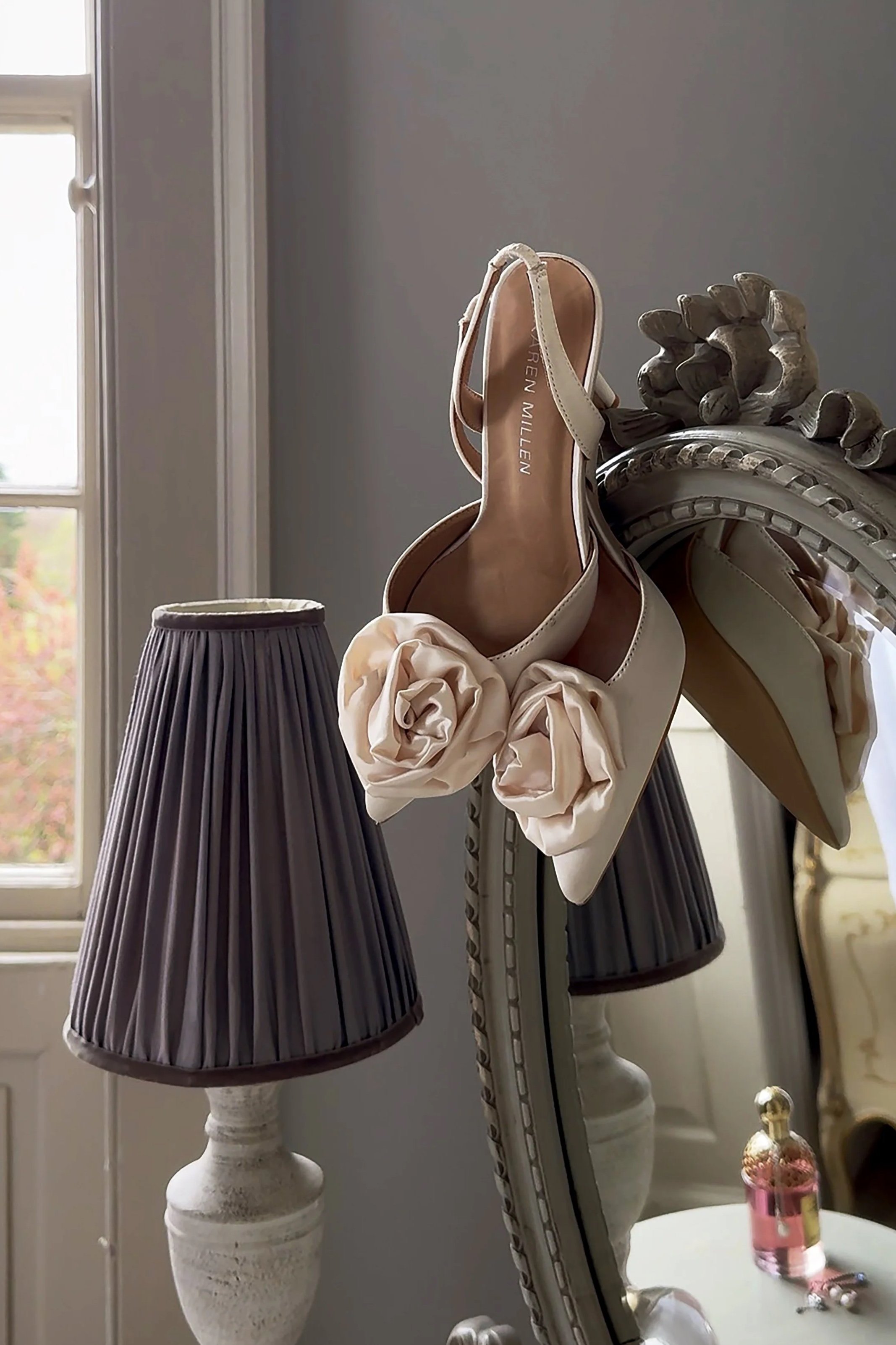 A pair of cream-colored high-heeled shoes with large fabric flowers on the toes, resting on a decorative mirror with black lampshades on nearby lamps, and a small bottle of pink perfume on a white table in the background.