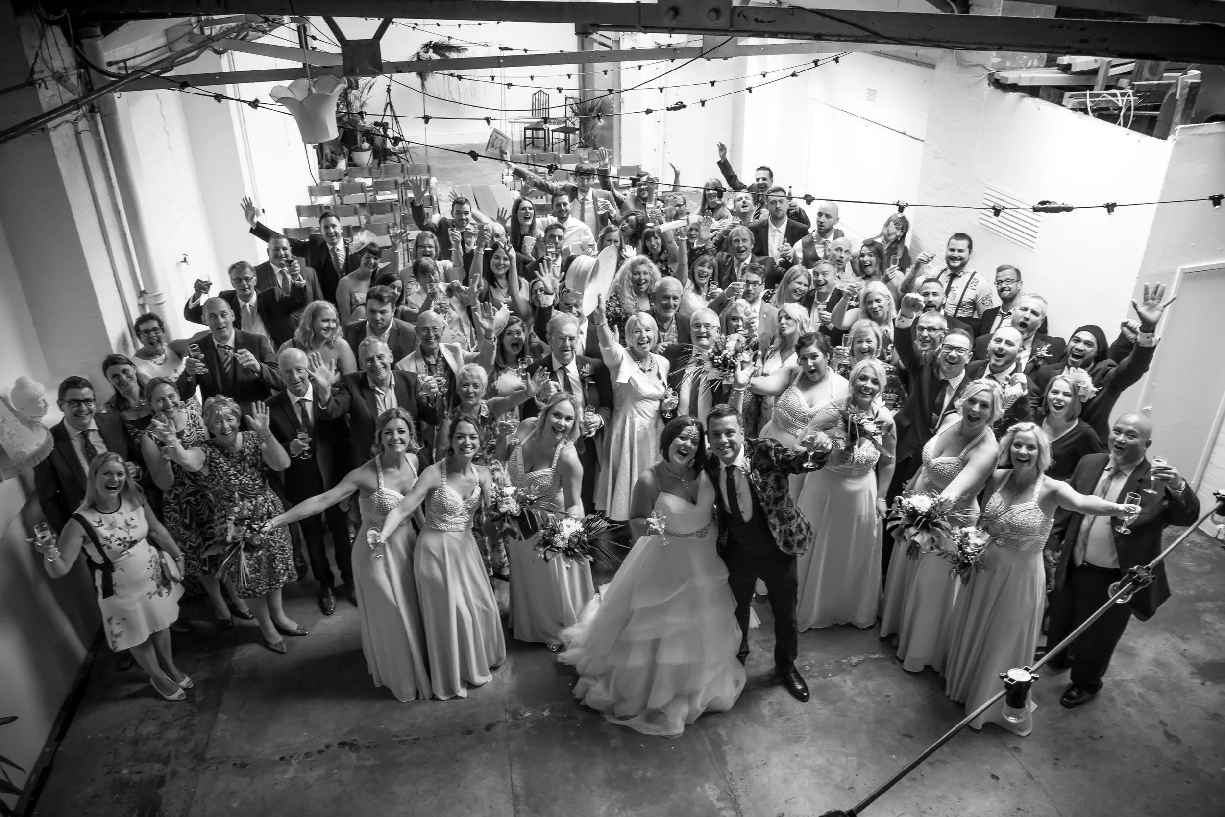 Black and white photo of a large group of people at a wedding celebration, including the bride and groom in the center, all smiling and waving.