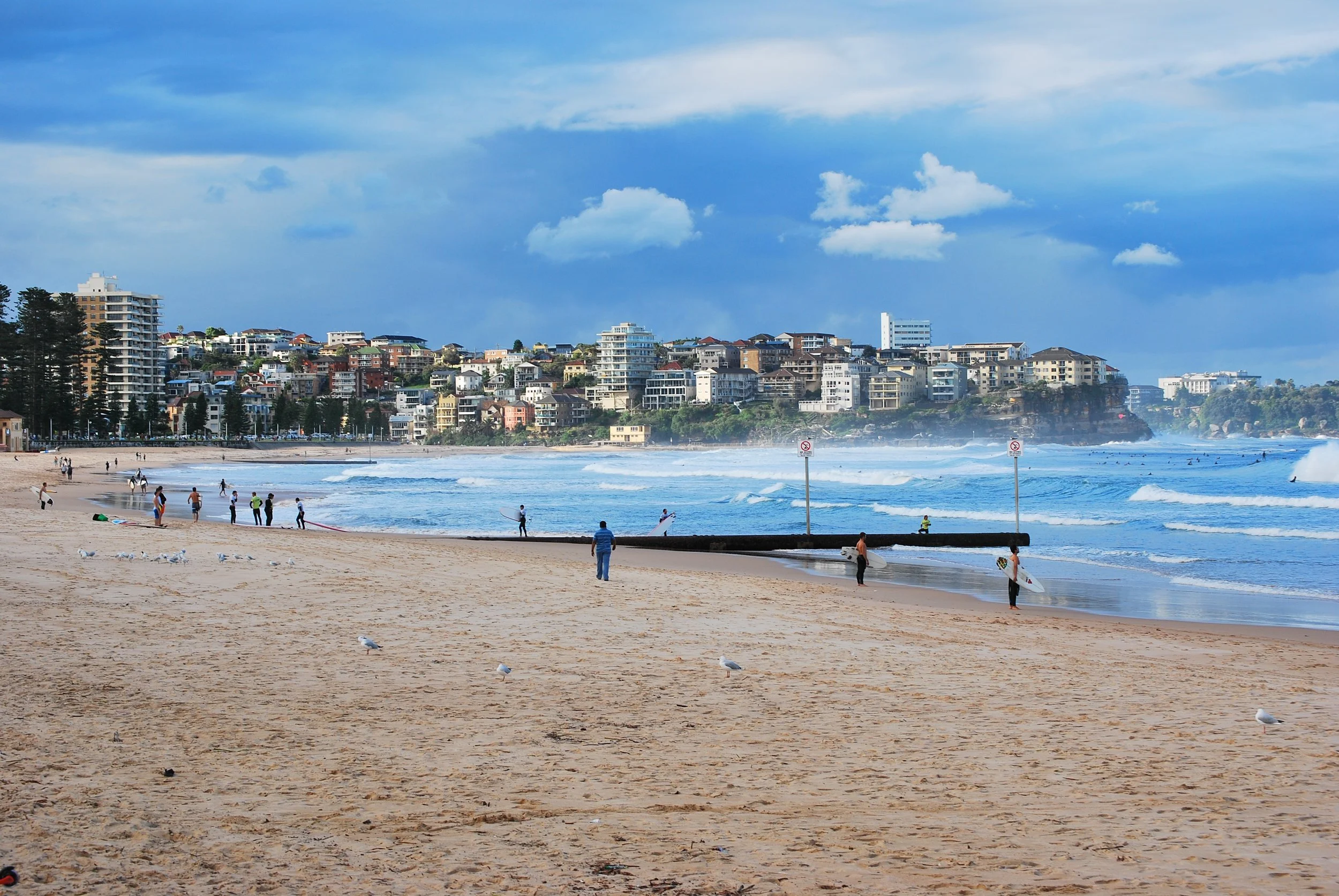 A sandy beach with people surfing and walking along the shore, with a cityscape of buildings on a hill in the background, under a partly cloudy sky.