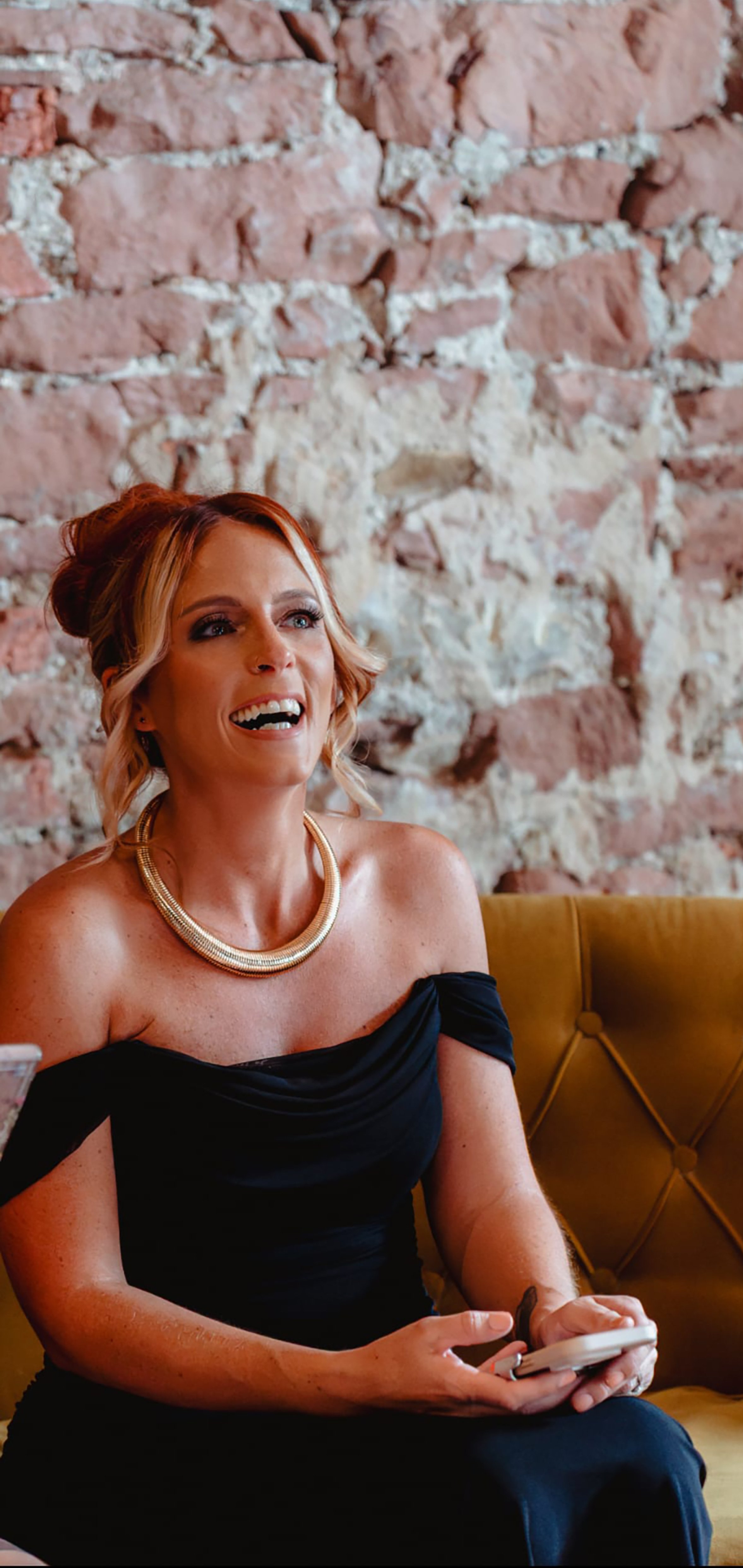 A woman with red hair styled in loose waves, wearing a black off-the-shoulder dress and a gold statement necklace, sitting on a yellow tufted sofa with a brick wall background, smiling while holding a smartphone.