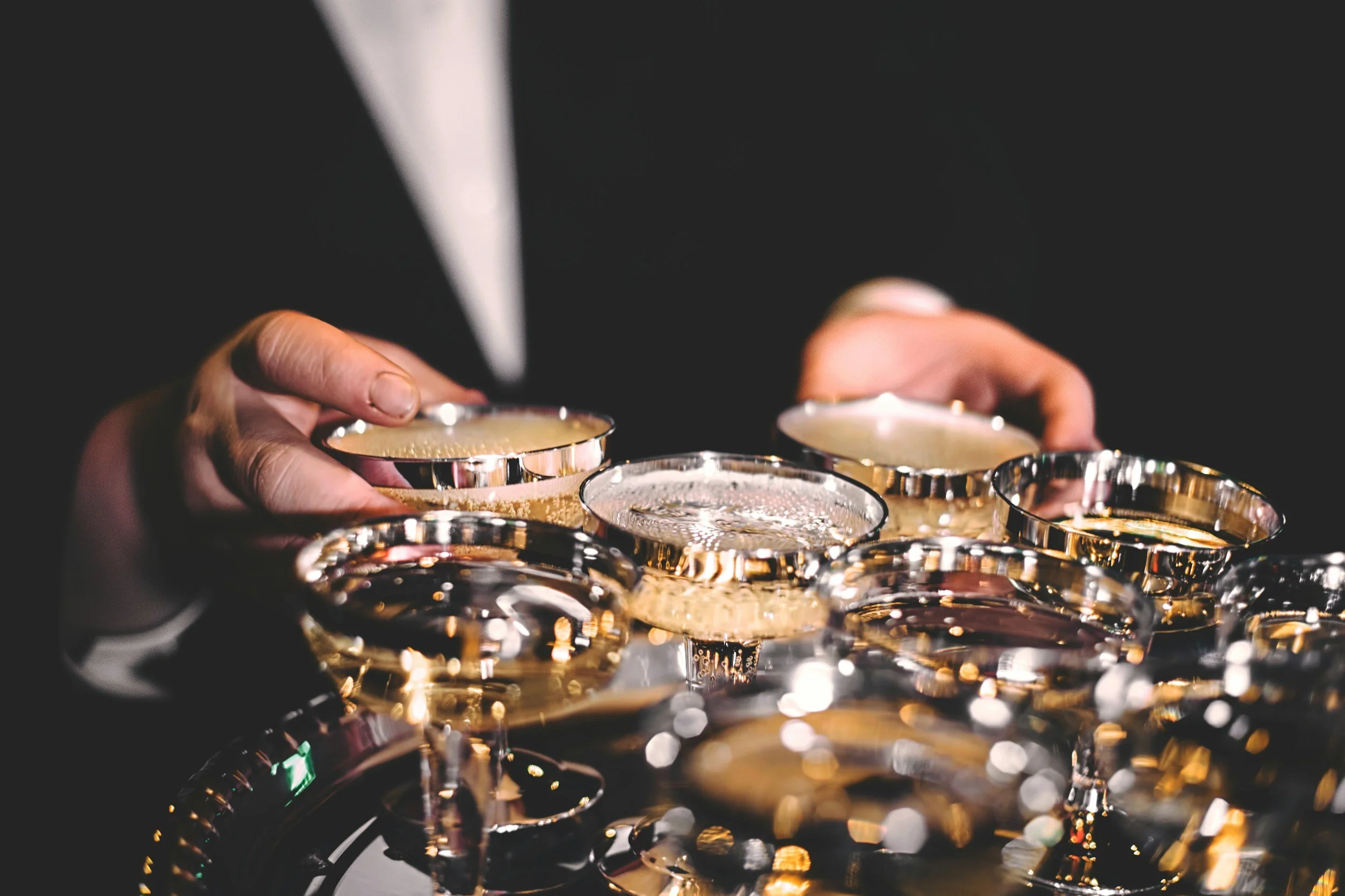 Close-up of a person's hands holding gold-colored cocktail glasses with bubbly drinks, with a blurred background.