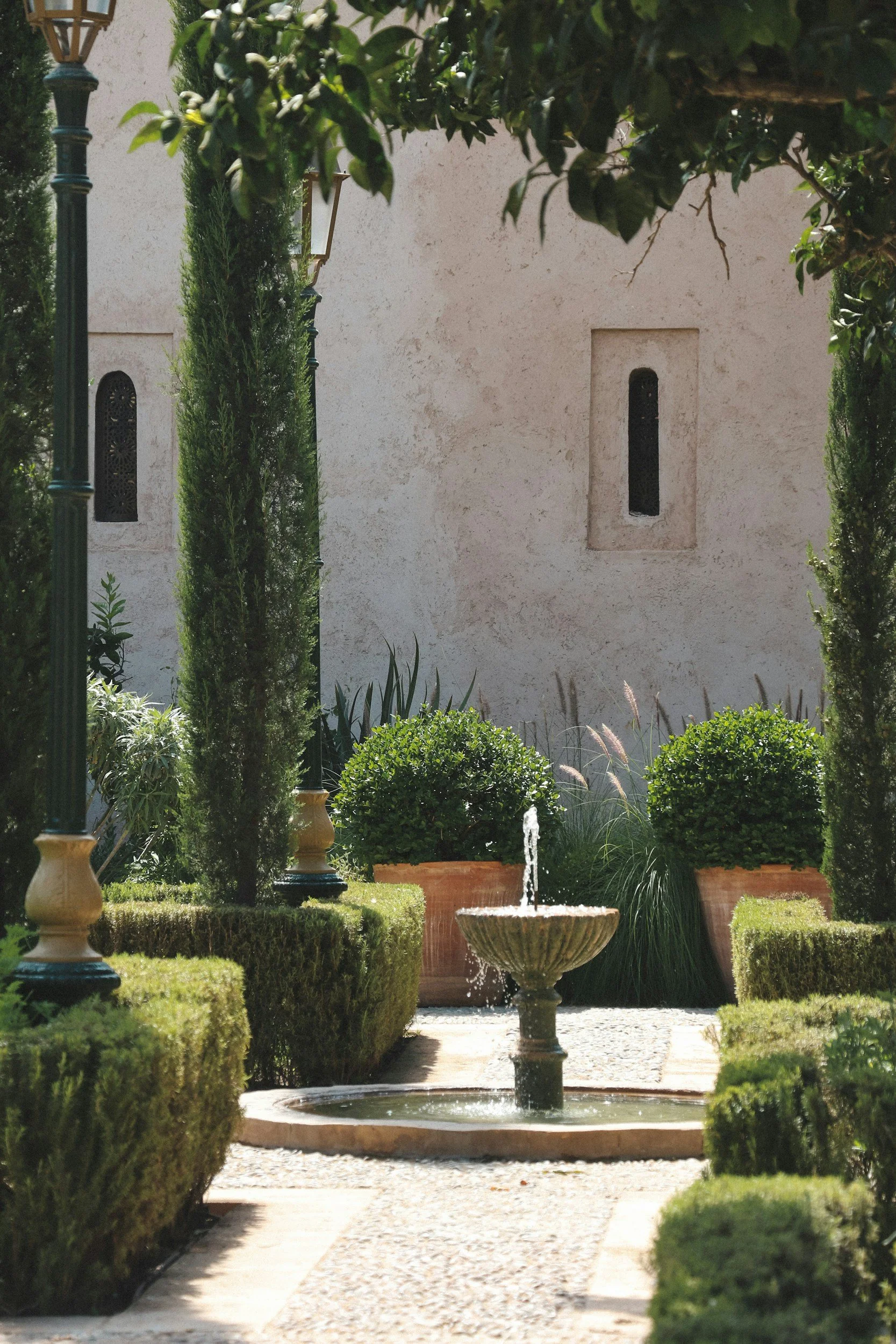 A serene garden with a small fountain surrounded by neatly trimmed bushes and potted plants, flanked by tall, slender trees, with a textured beige wall and small narrow windows in the background.