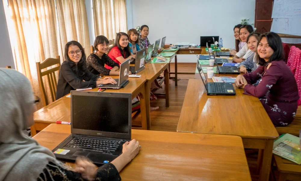 Group of women sitting around a large wooden conference table at YMCA headquarters in Yangon Myanmar with laptops provided by the Jared Prescott Trust during a meeting in a bright room.