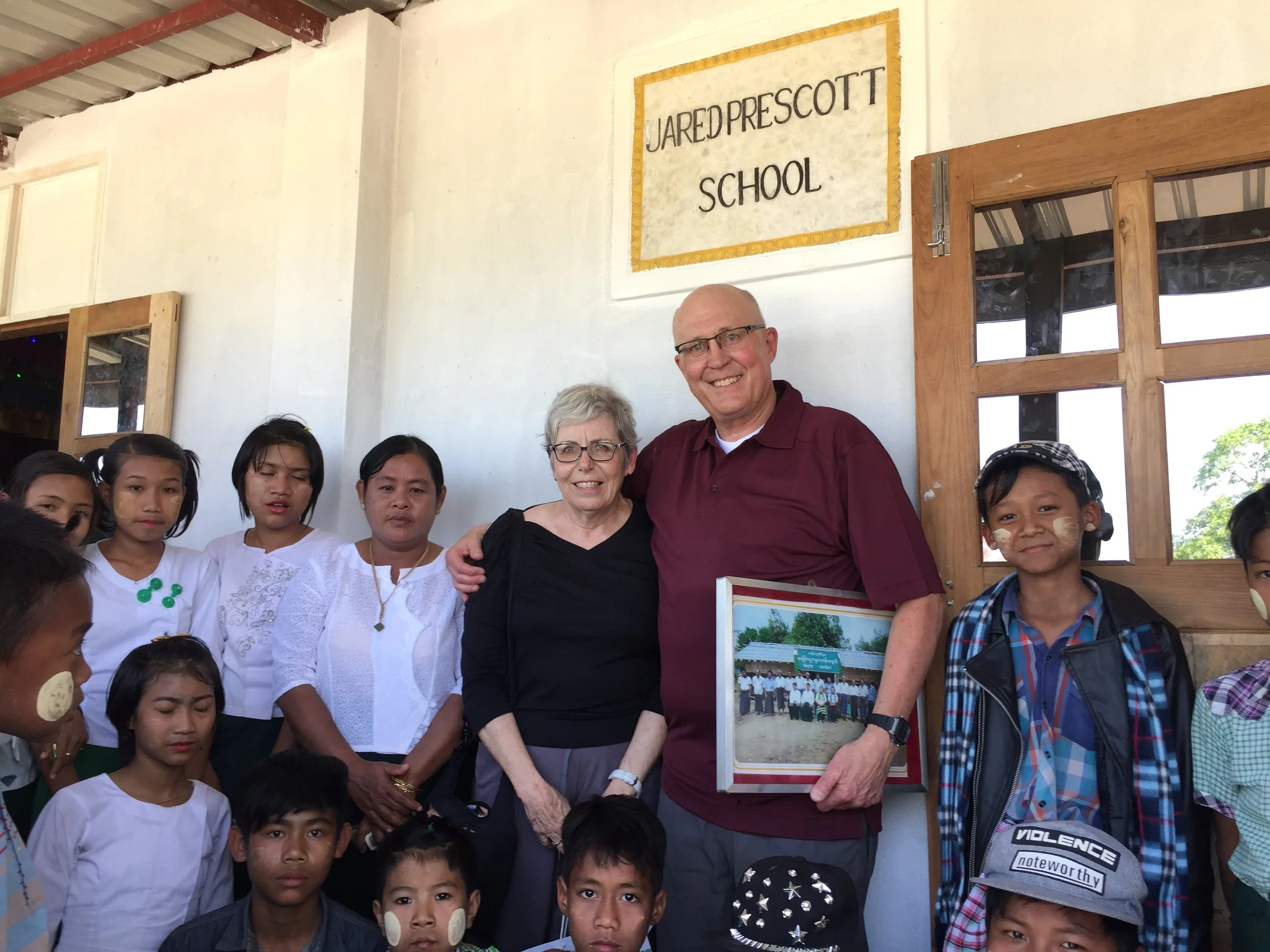 Group of children and two adults, a man and a woman, standing outside Jared Prescott School building with a sign above the door. The children are wearing traditional clothing with face paint and some are looking at the camera.