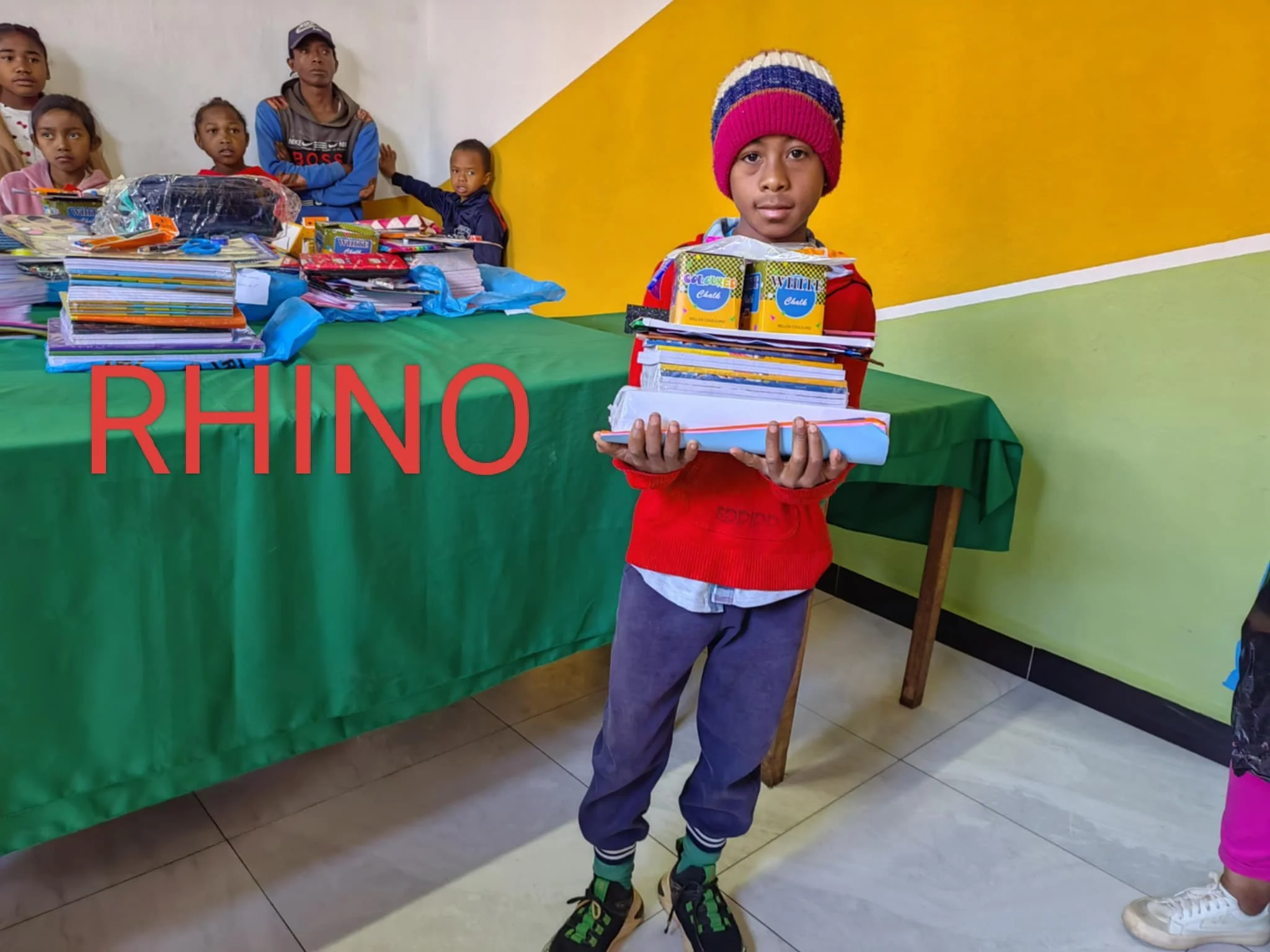 A young boy standing in front of a table with books and school supplies, holding a stack of notebooks and folders, wearing a red sweater, blue pants, and a colorful knit beanie. Several children are in the background, some looking at the camera, with a colorful yellow and green wall behind them.