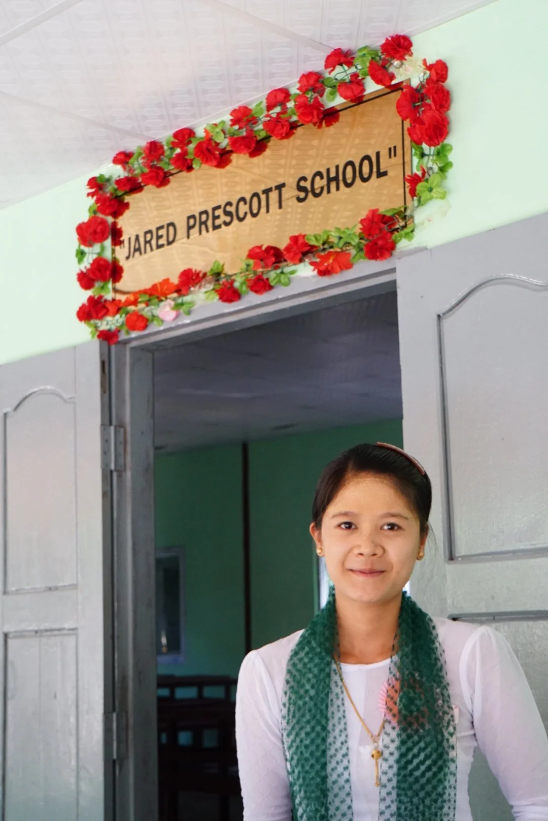 A young teacher standing in front of the entrance to a new Jared Prescott School, which has a sign with the school's name decorated with red and green artificial flowers.